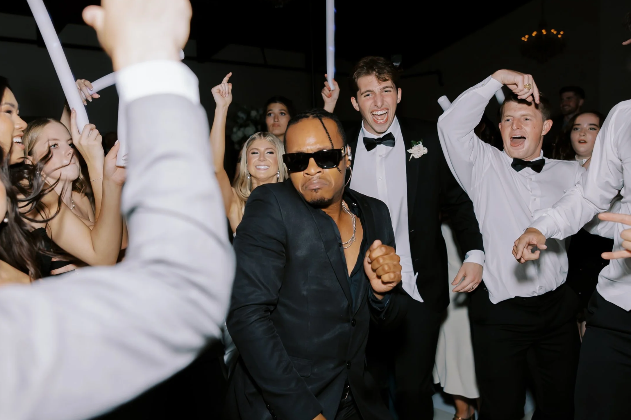 People dancing and celebrating at a party, with a man wearing sunglasses and a woman in a tuxedo in the foreground.