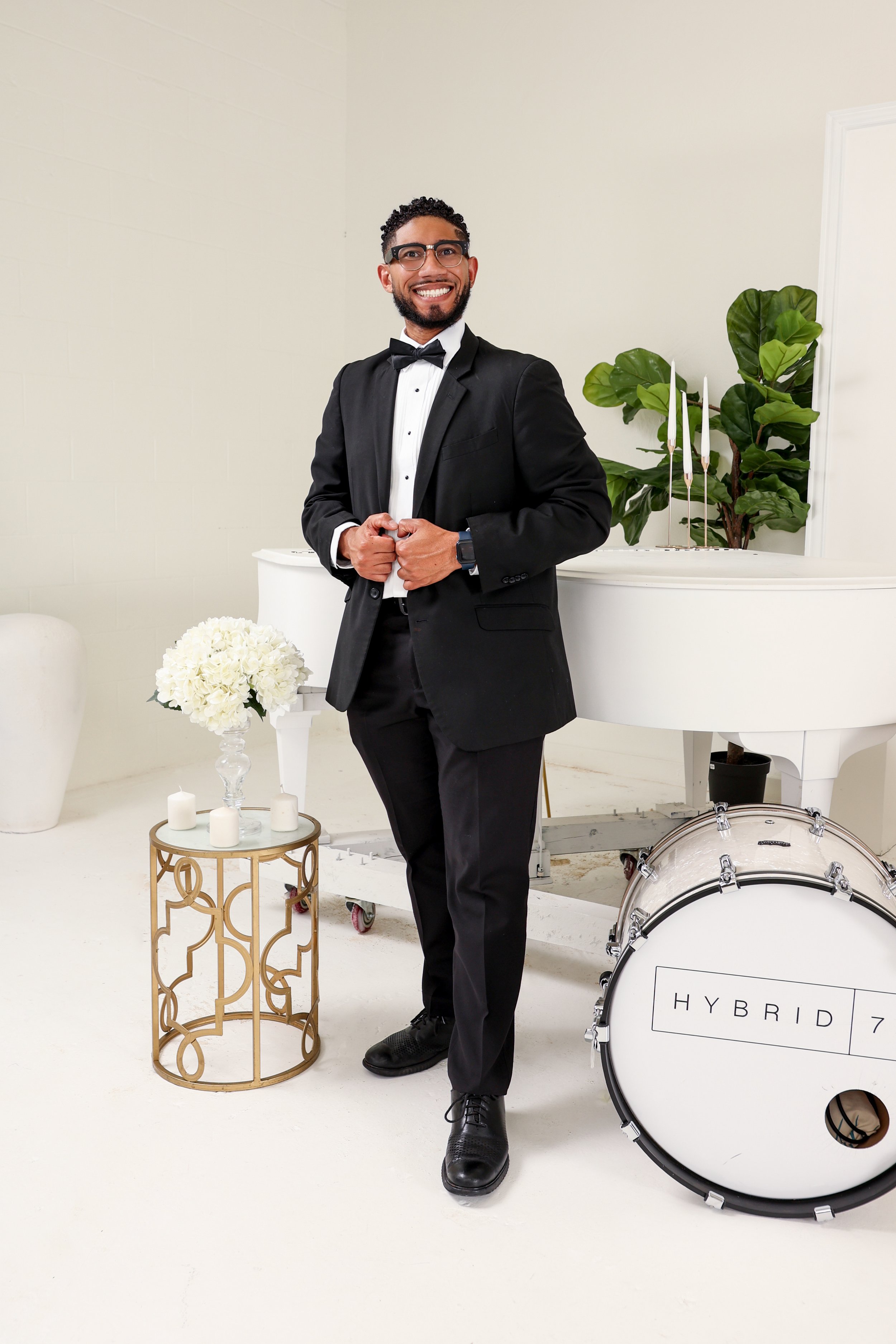 Man in black tuxedo with bow tie standing in a white room with a flower arrangement, a drum labeled 'HYBRID 7', and a white piano.