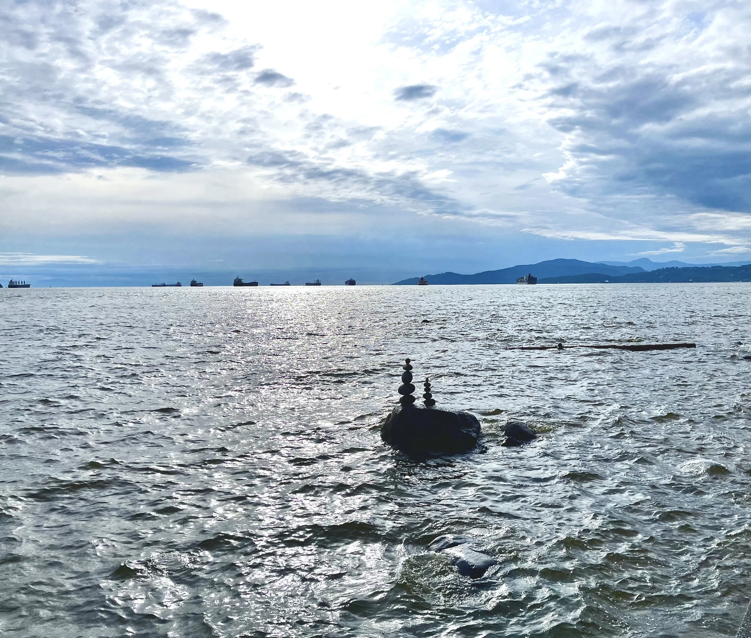 Ocean view with a cloudy sky, several ships on the horizon, and stacked stones balanced on a rock in the water in the foreground.