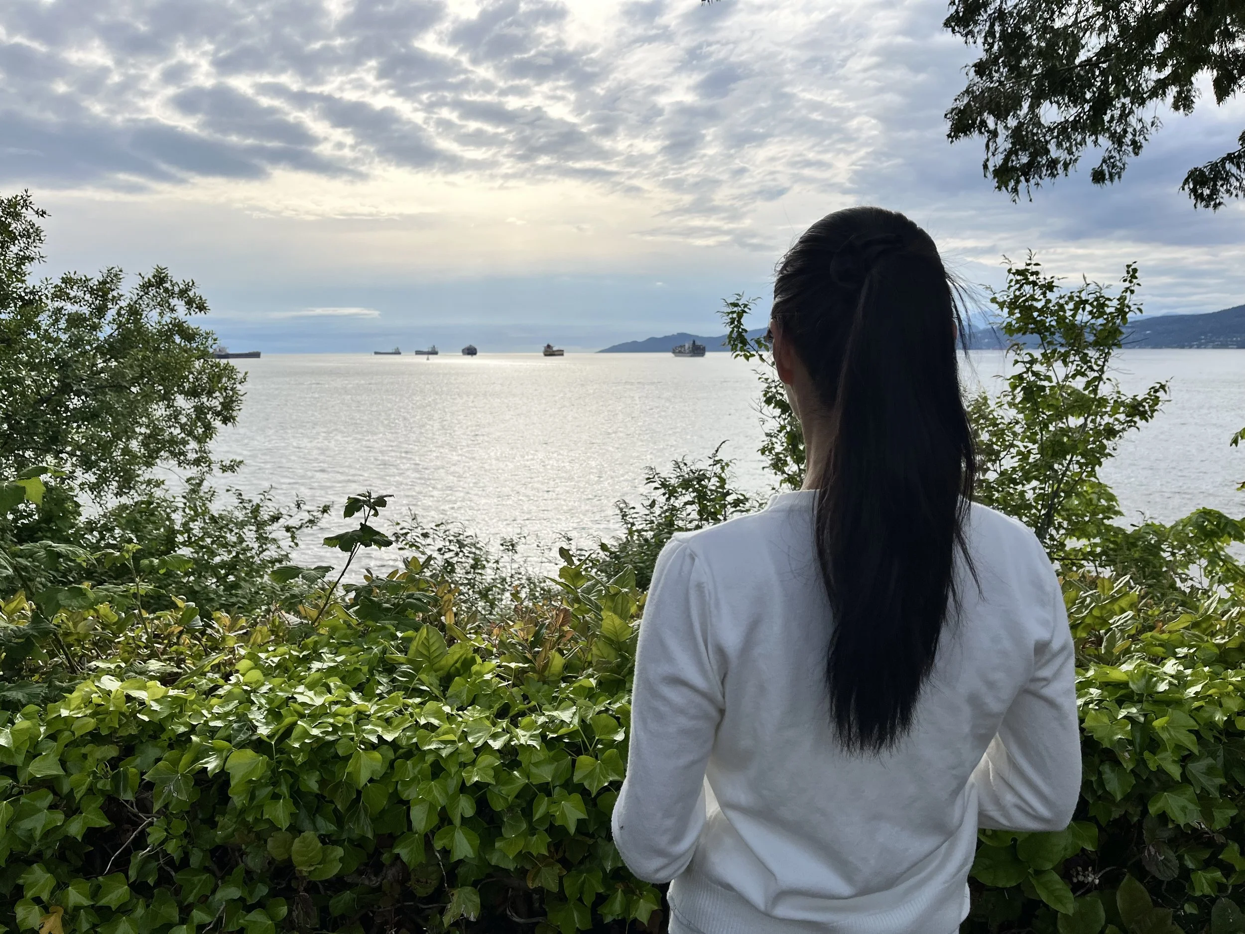 A woman with long dark hair, wearing a white top, standing outdoors among green bushes, looking at a body of water with ships and a distant shoreline under a cloudy sky.