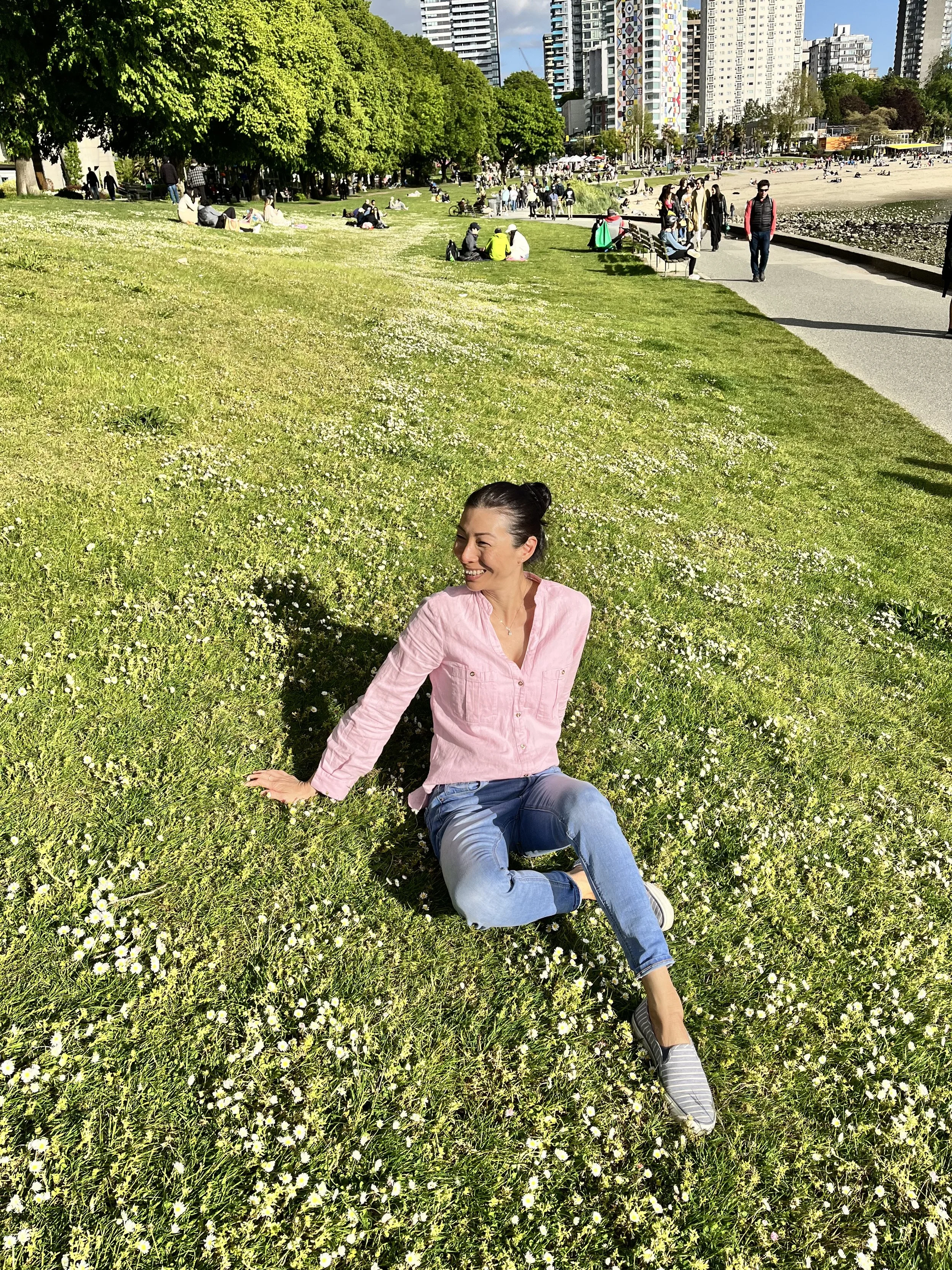 Woman sitting on grass surrounded by white flowers in a park with trees, people, and city buildings in the background.