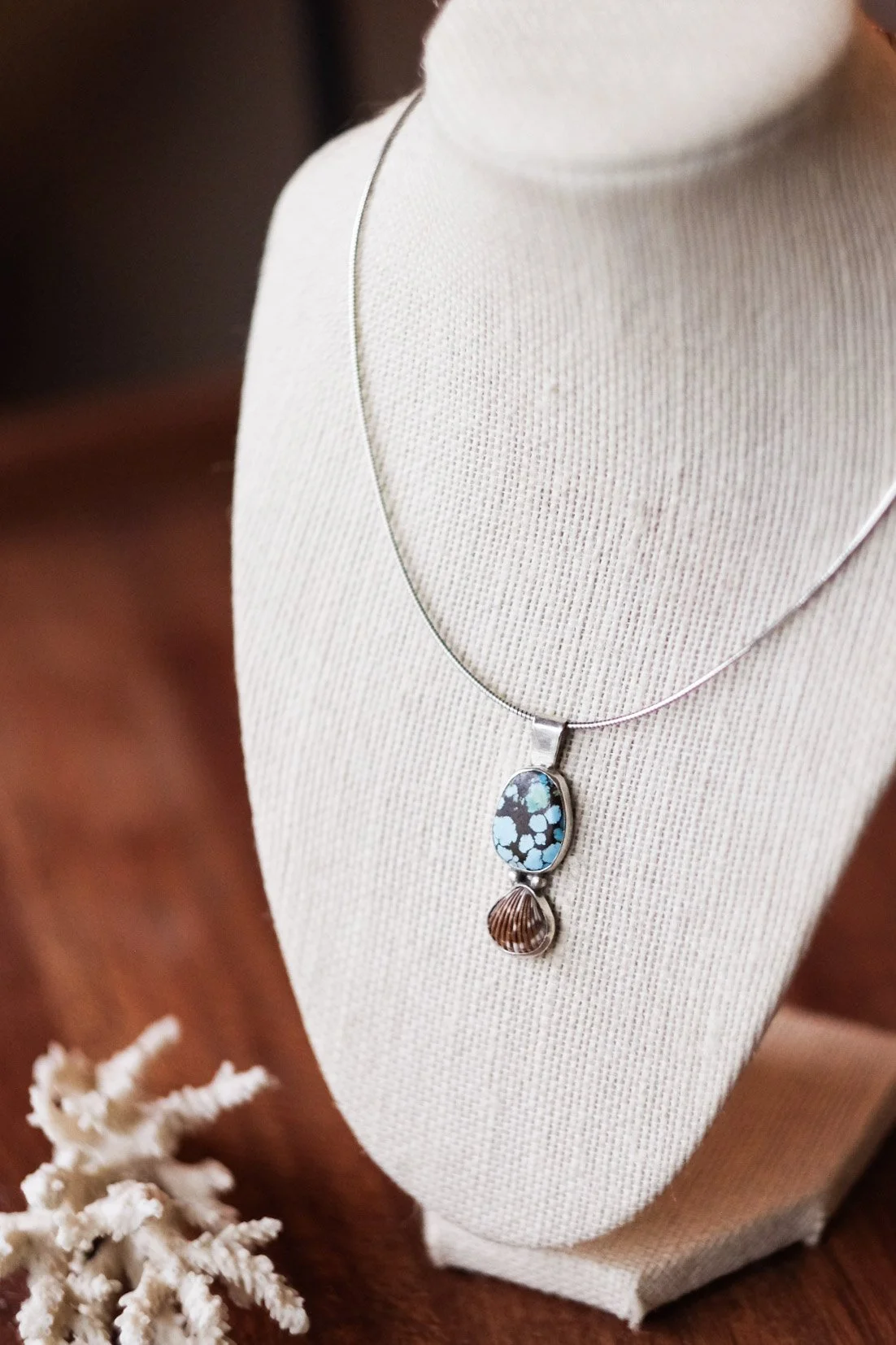 A necklace with a large oval turquoise and black stone pendant and a smaller scalloped shell pendant displayed on a jewelry bust.