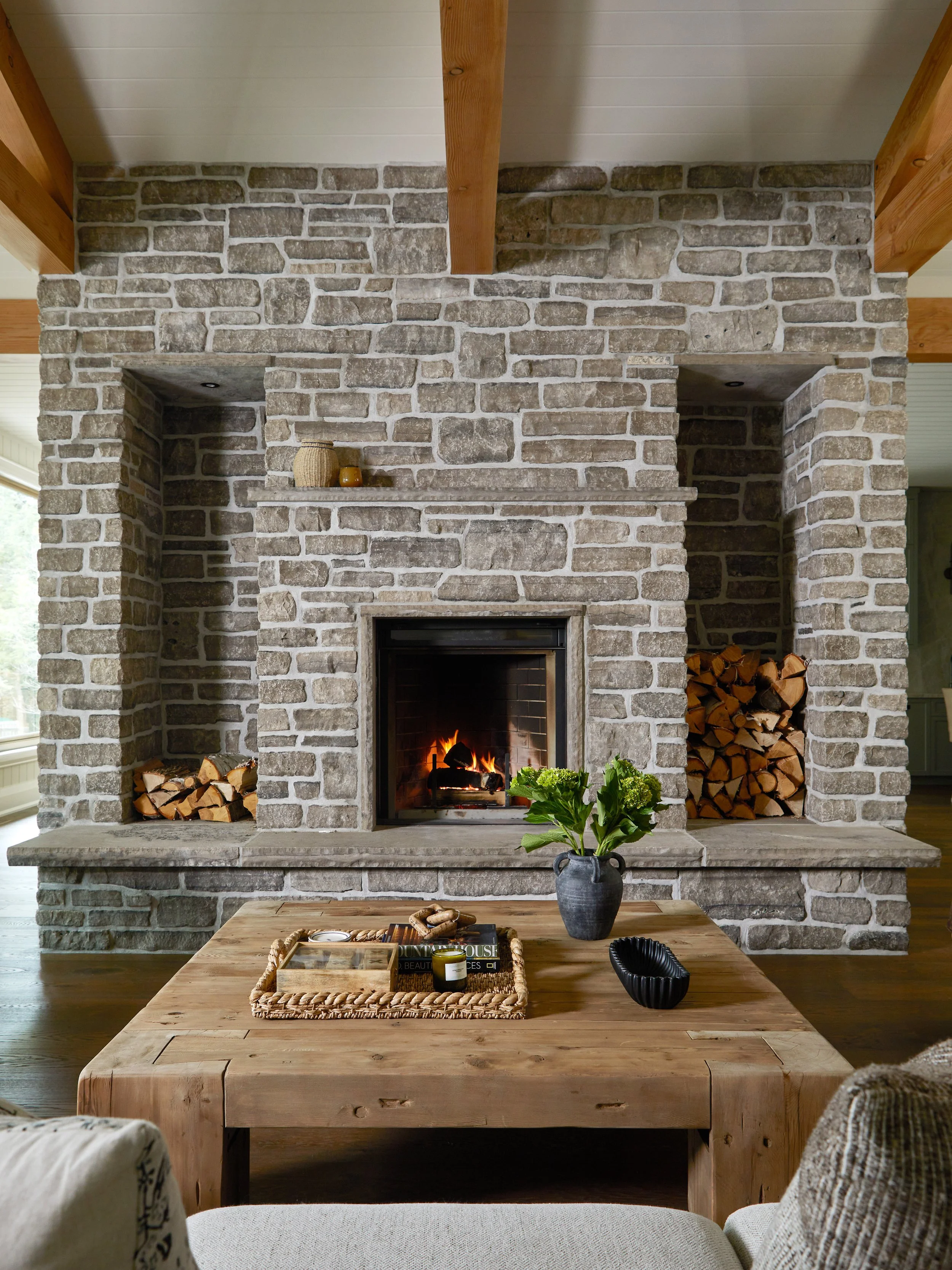 Living room with a rustic stone fireplace, wood logs stored on the sides, a wooden coffee table with decorative items and a pitcher of green flowers, and cozy seating.