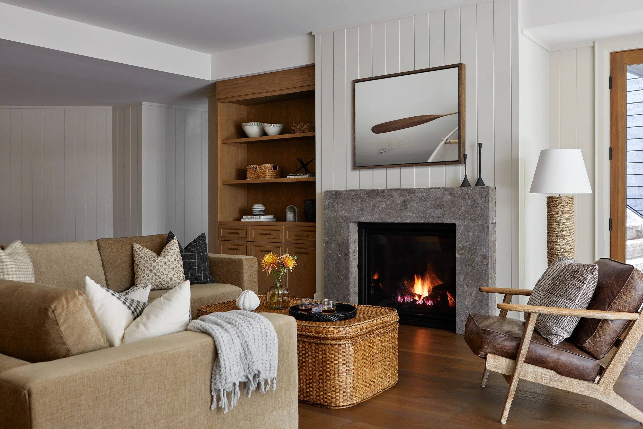Living room with beige sofa, brown leather armchair, wooden coffee table, fireplace with a fire, built-in wooden shelves, wall-mounted mirror, floor lamp, and window with wooden trim