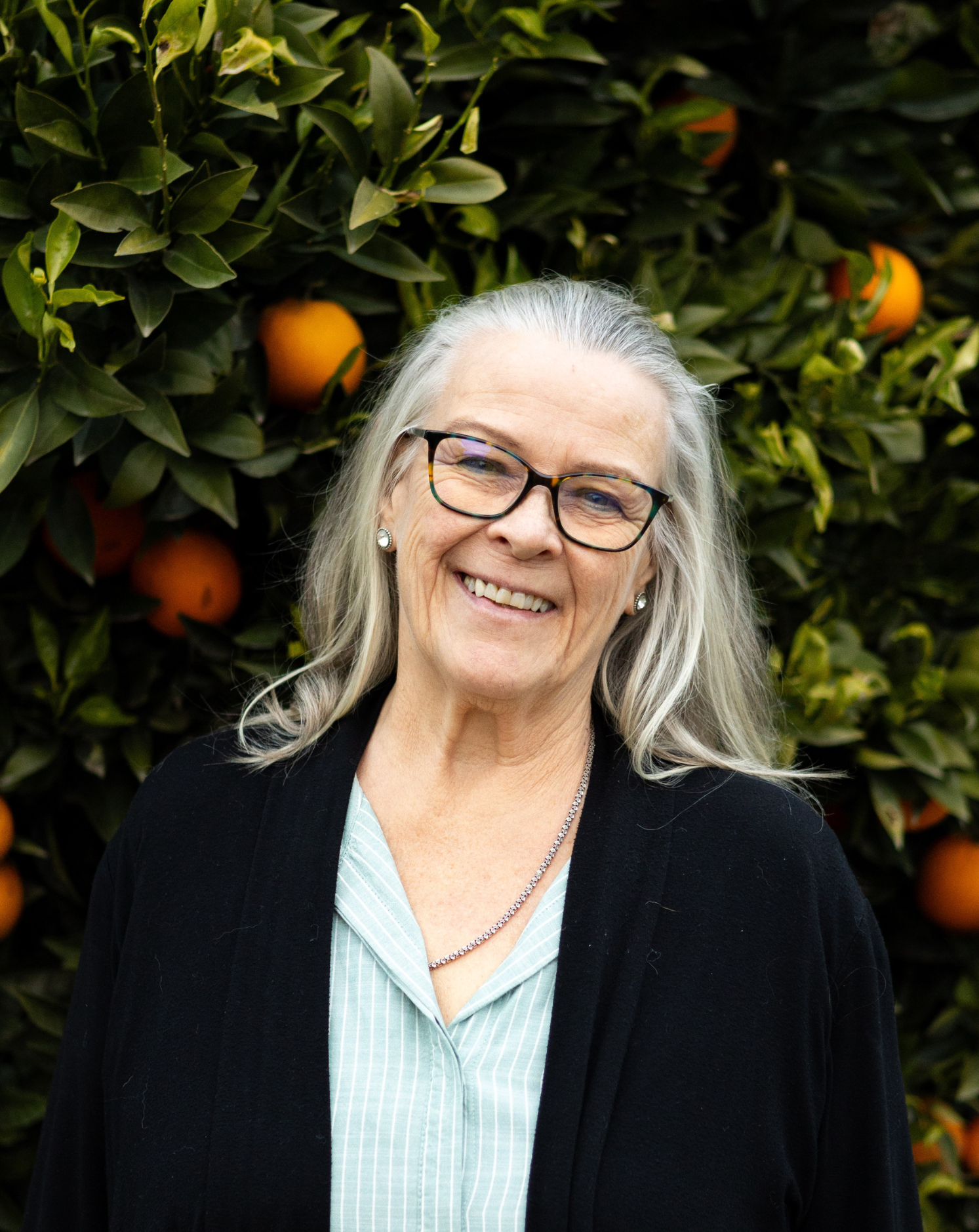 Smiling elderly woman with long gray hair and glasses standing in front of an orange tree.