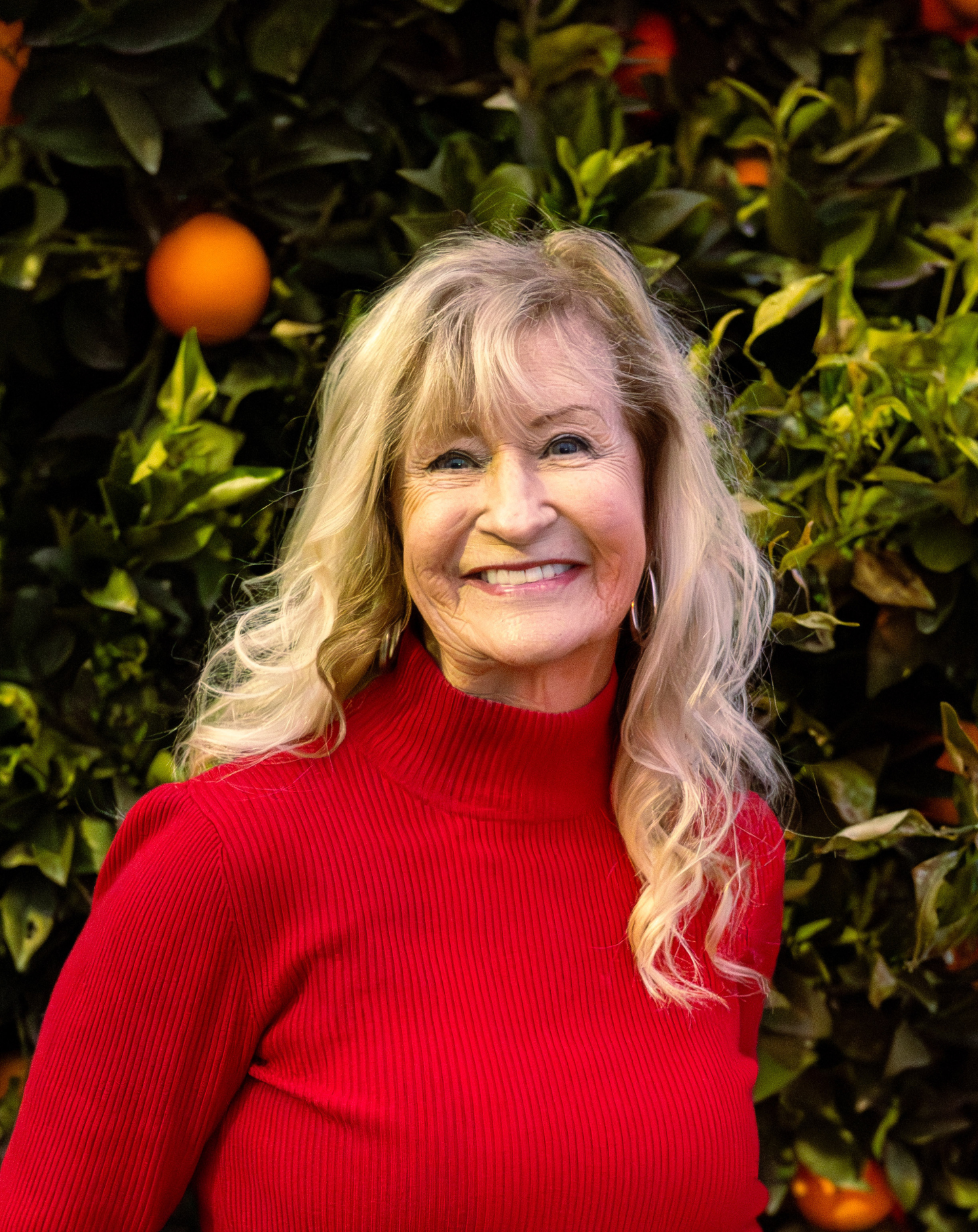 A smiling woman with long blonde hair wearing a red turtleneck sweater standing in front of a backdrop of green leaves and orange fruit.