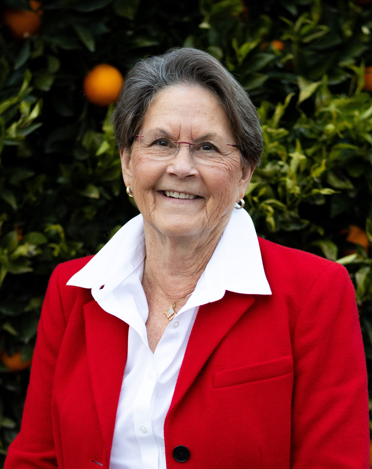 An older woman in a red blazer and white shirt standing outdoors with an orange tree in the background.
