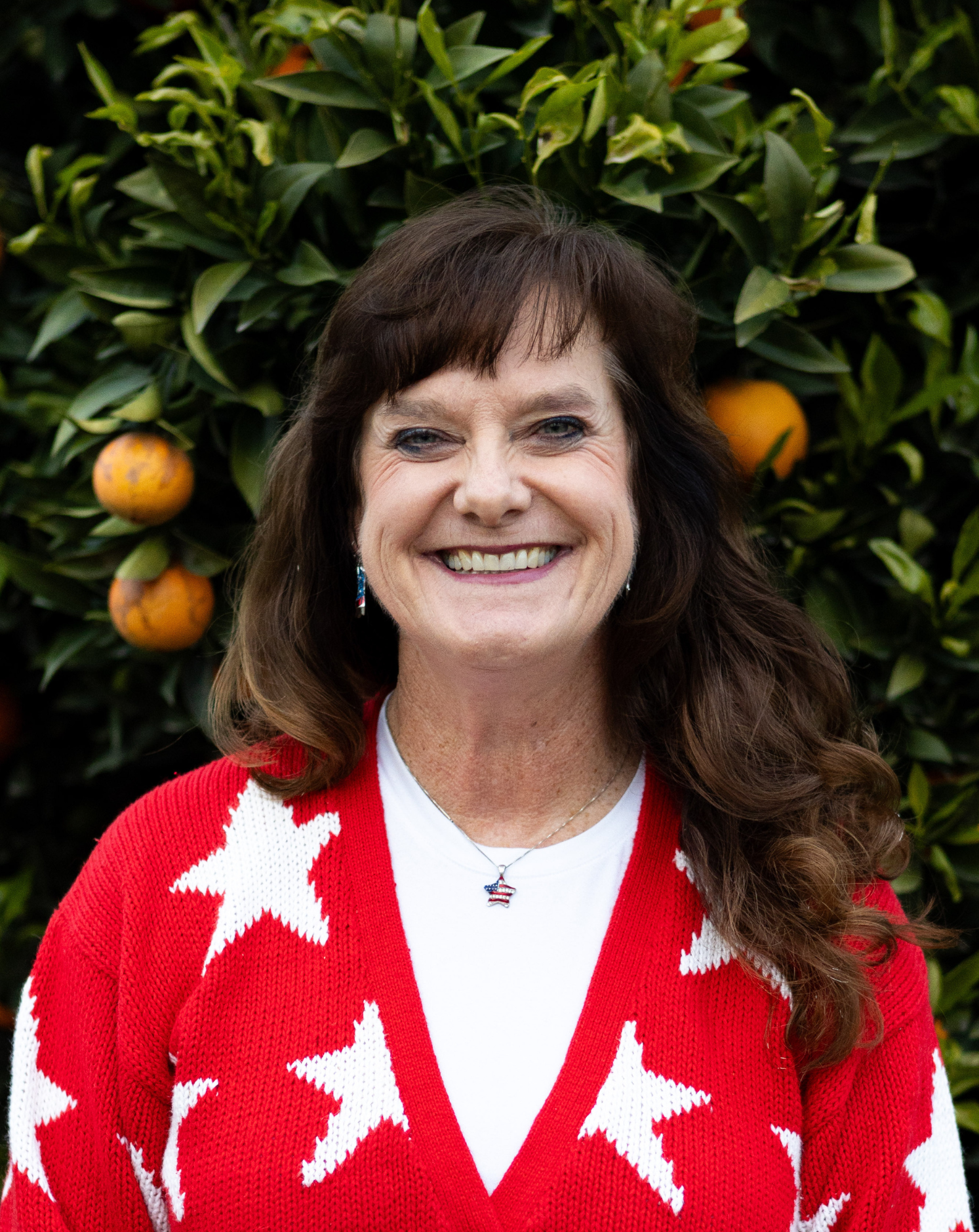 A smiling woman with brown wavy hair, wearing a red sweater with white stars, a white shirt, and star-shaped earrings, stands in front of an orange tree with ripe oranges.