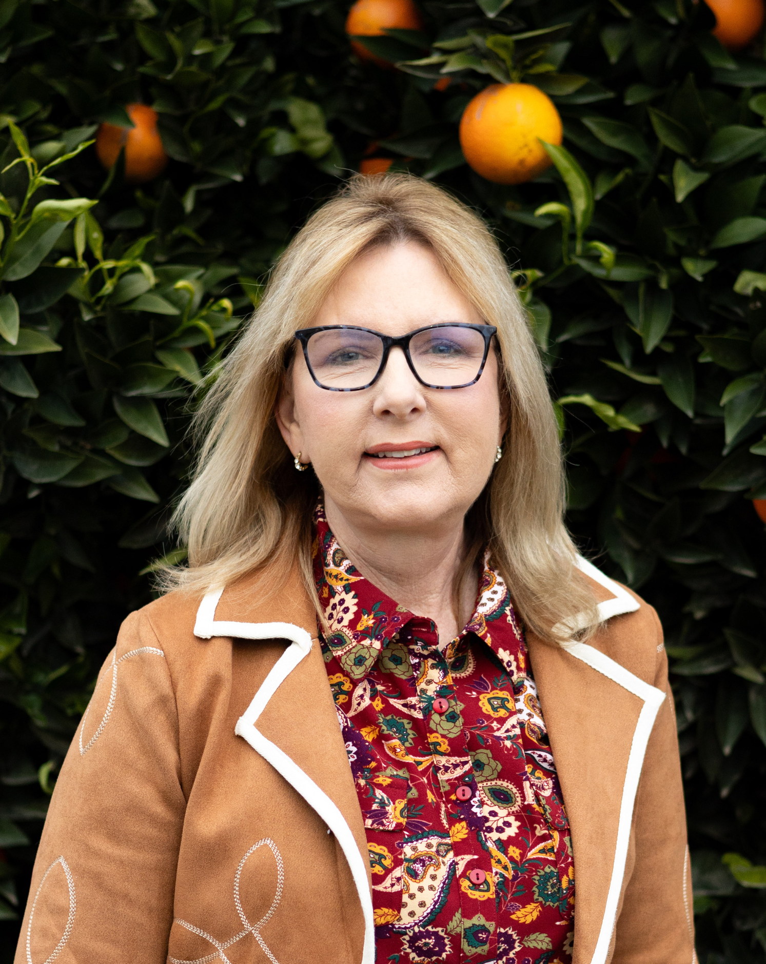 A woman with blonde hair wearing glasses, a patterned red blouse, and a tan jacket with white trim, standing in front of a hedge with oranges.