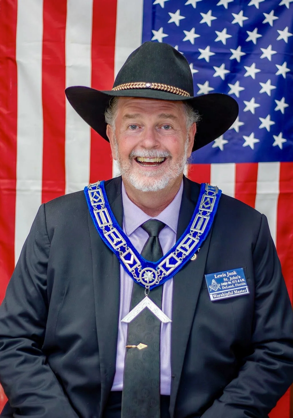 A smiling man wearing a black suit, purple shirt, and gray tie, decorated with a Masonic collar and chain, standing in front of an American flag.