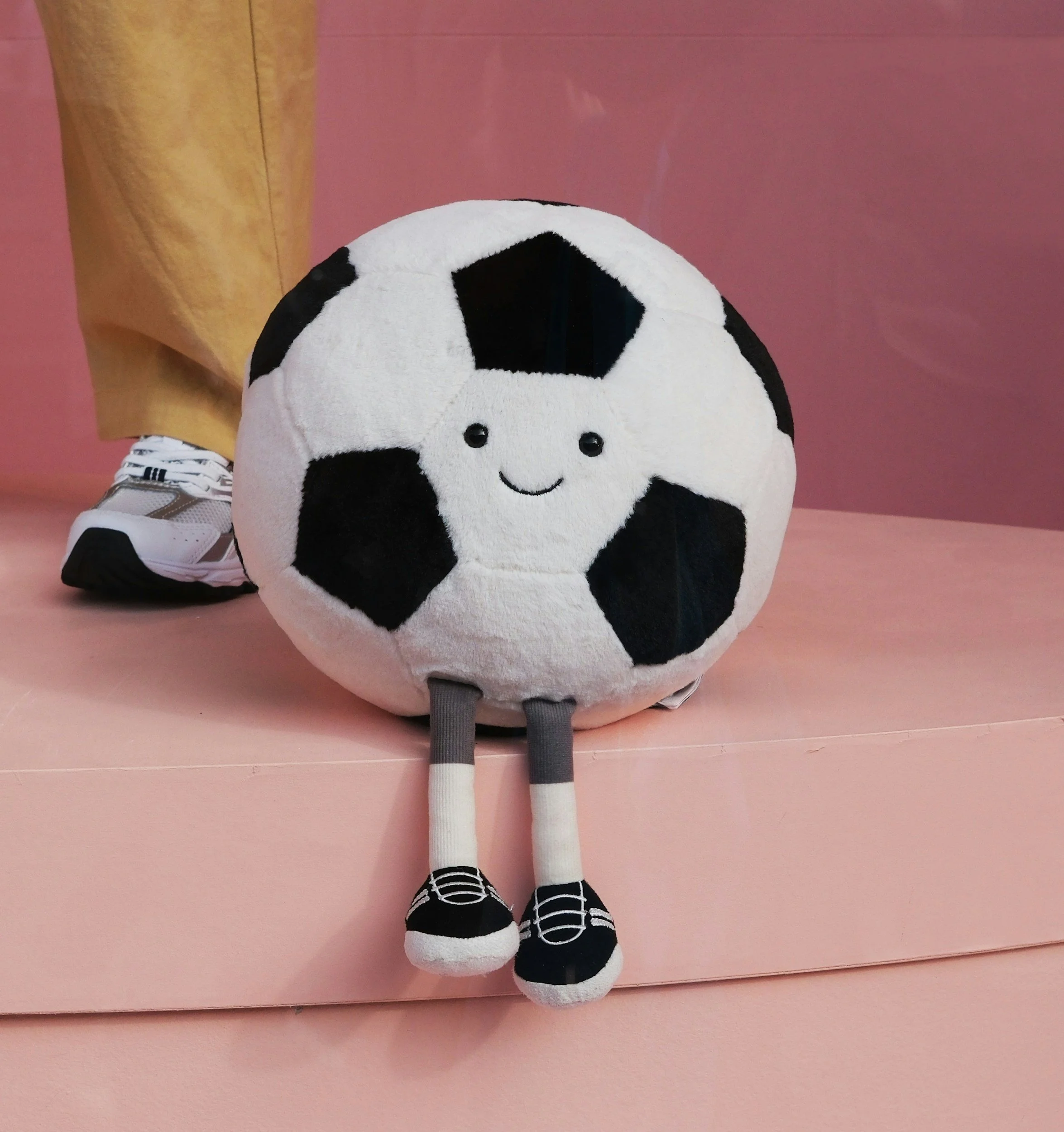 A plush soccer ball with a smiling face and wearing cartoon sneakers, sitting on a pink surface.