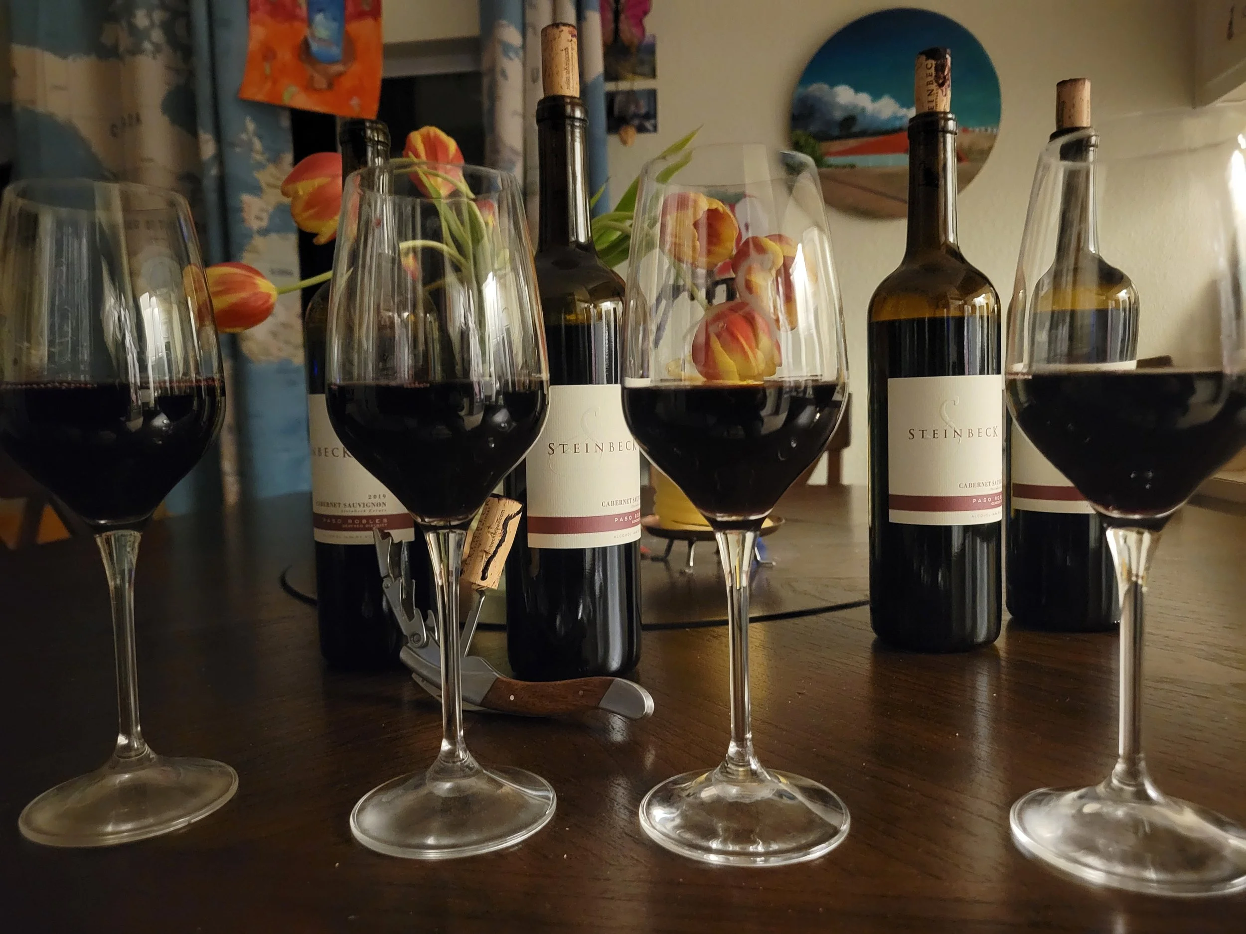 Four glasses of red wine on a wooden table with three bottles of wine and a bouquet of tulips in the background.