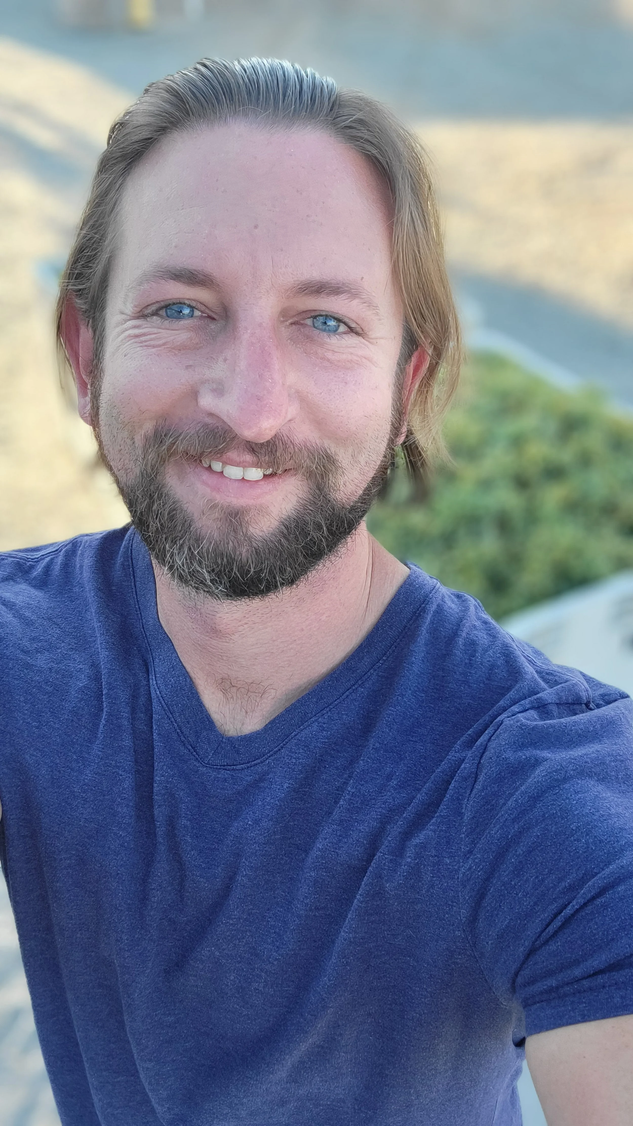 A smiling man with blue eyes, brown hair, and a beard, taking a selfie outdoors, with blurred greenery and a road in the background.