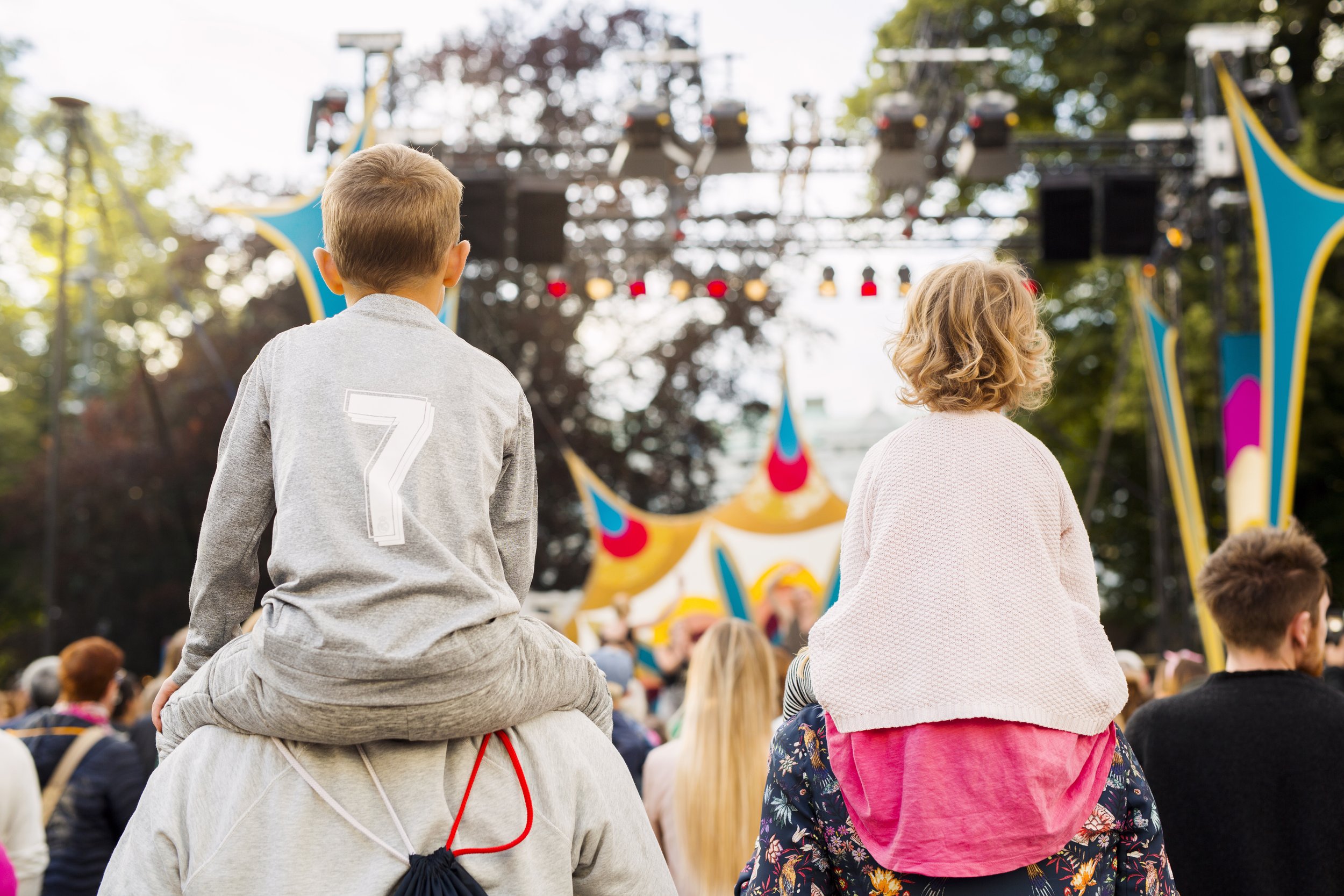 Children sitting outdoors at a festival or concert, facing a stage with colorful decorations and lighting, surrounded by trees.
