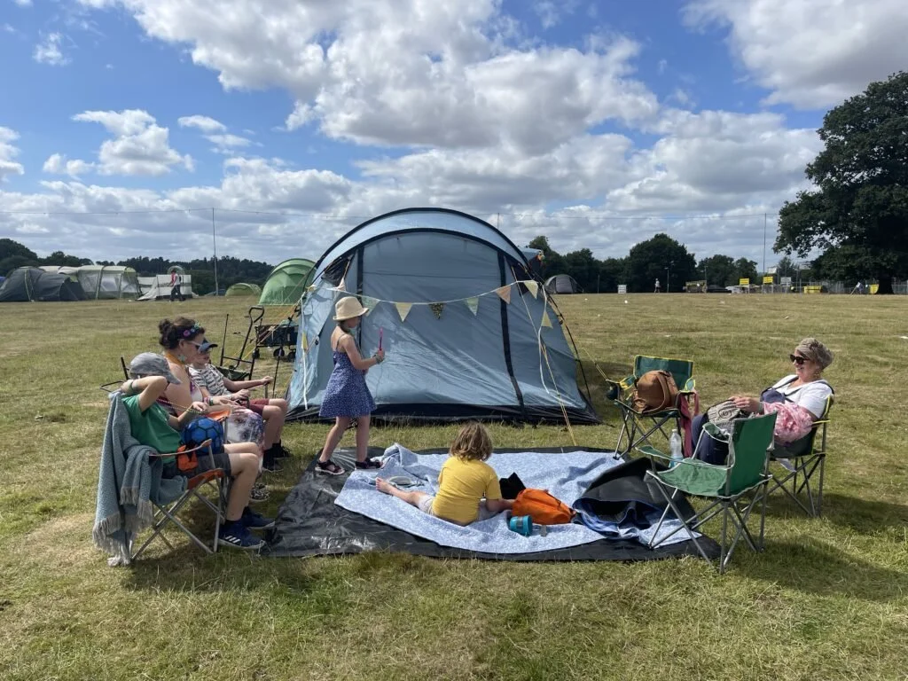 A group of children and an adult sitting on camping chairs and a blanket on a grassy field near a blue tent, with other tents and trees in the background under a partly cloudy sky.