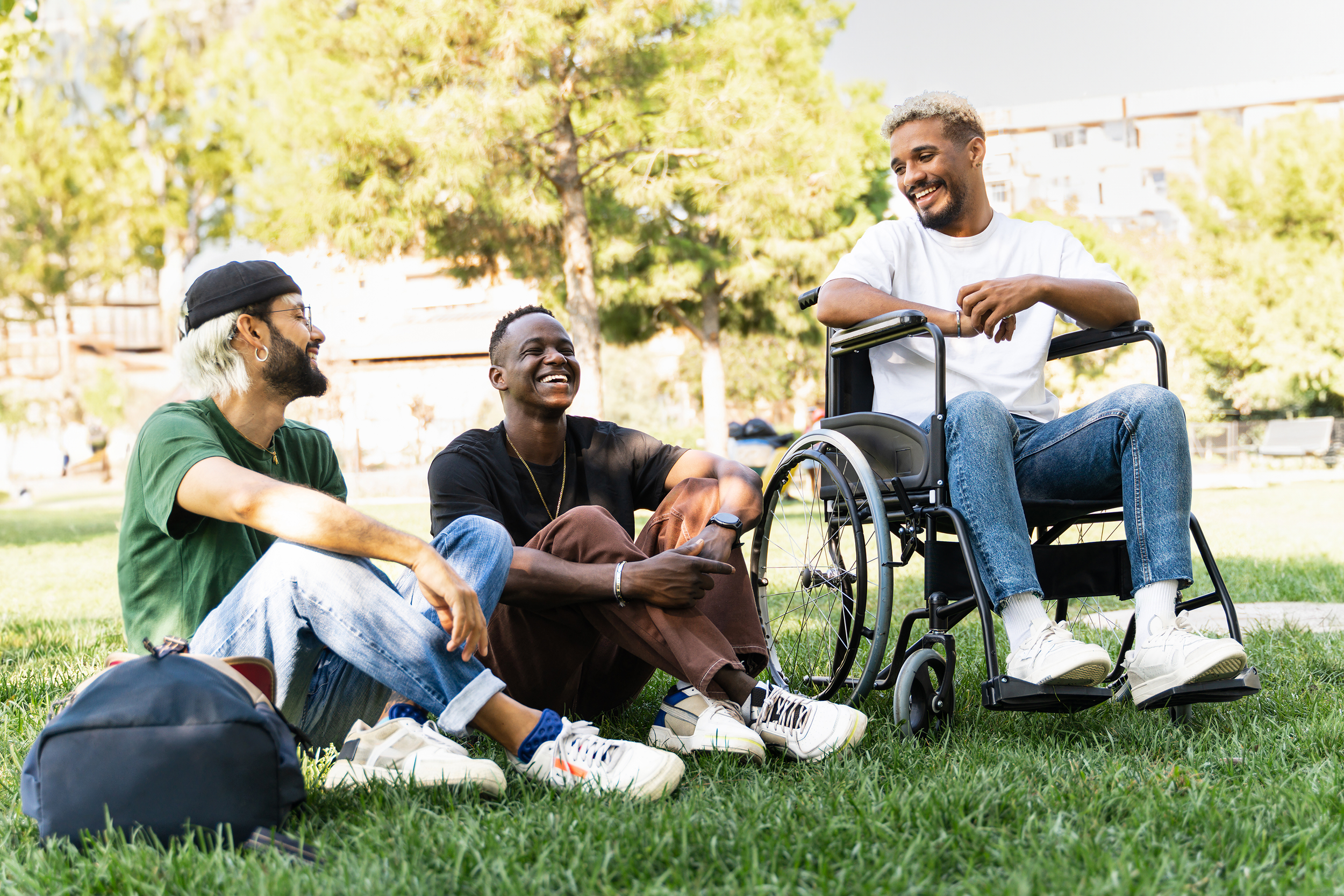 Three young men sitting on grass in a park, laughing and chatting, with trees and buildings in the background. One man is in a wheelchair.