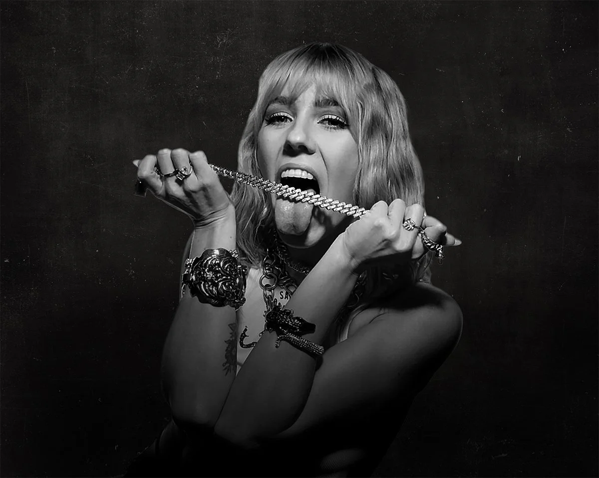 A woman with blonde hair holding a chain necklace with her tongue out.