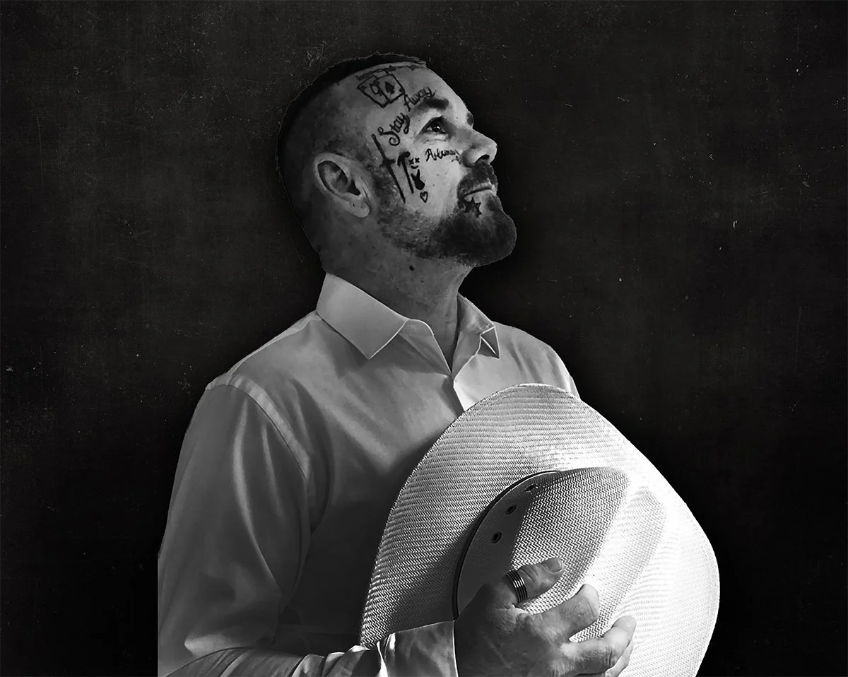A man looking upwards, holding a hat, with tattoos on his face and head, wearing a white button-up shirt, standing against a dark background.