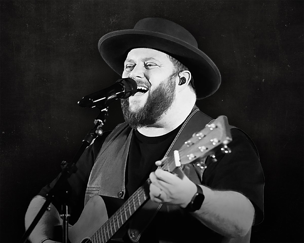 Man with a beard, sunglasses, and cap singing into a microphone while playing an acoustic guitar against a black background.