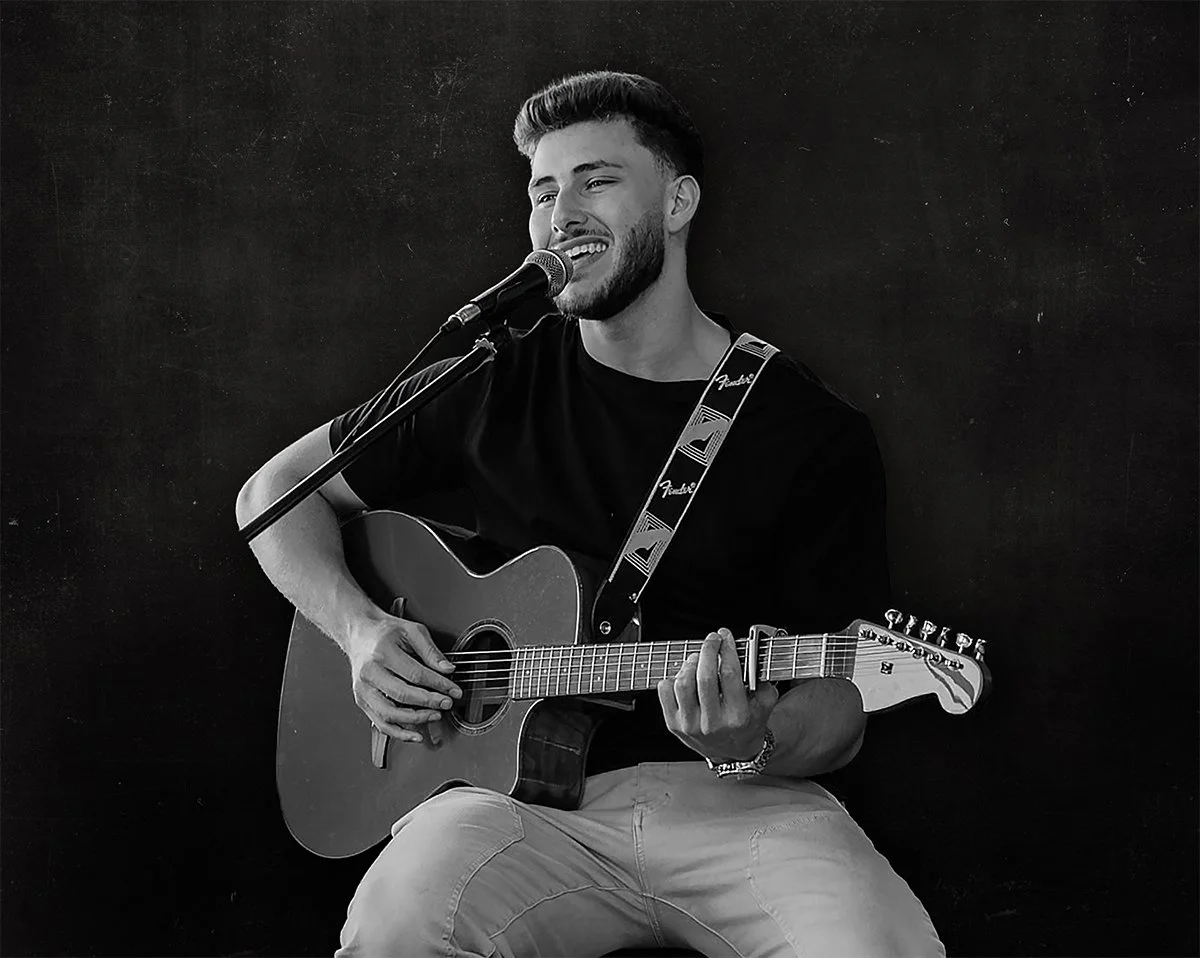 A young man with a beard and short hair, wearing a black t-shirt and beige pants, is sitting and playing a blue acoustic guitar while singing into a microphone.