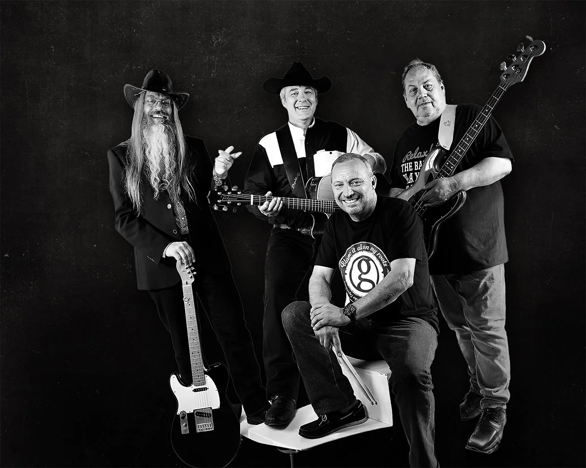 Four men posing with musical instruments against a colorful background. One man is sitting on a white block, while the others are standing, holding guitars and a bass guitar. The group appears to be a band.