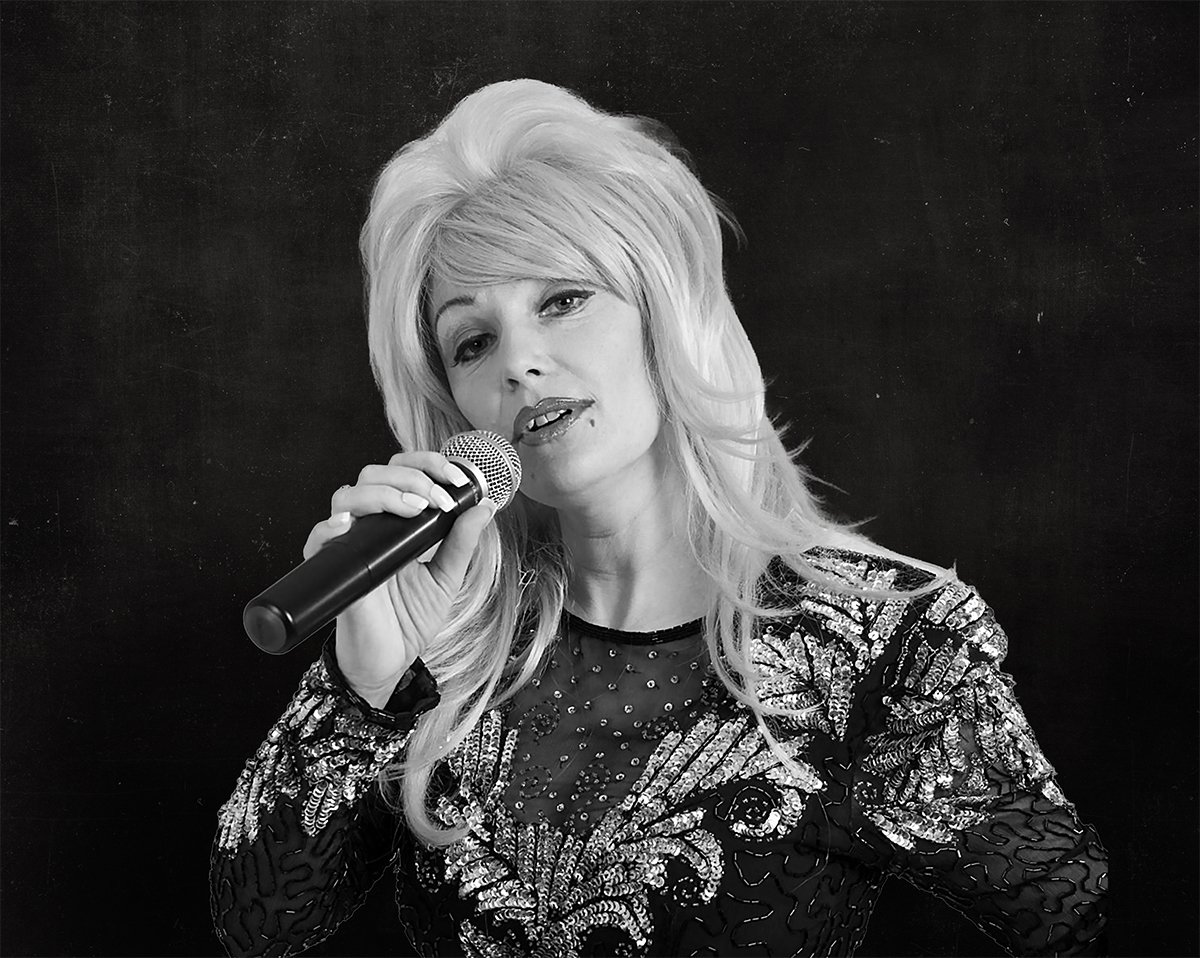 A smiling woman with blonde hair holding an acoustic guitar in front of a plain background.