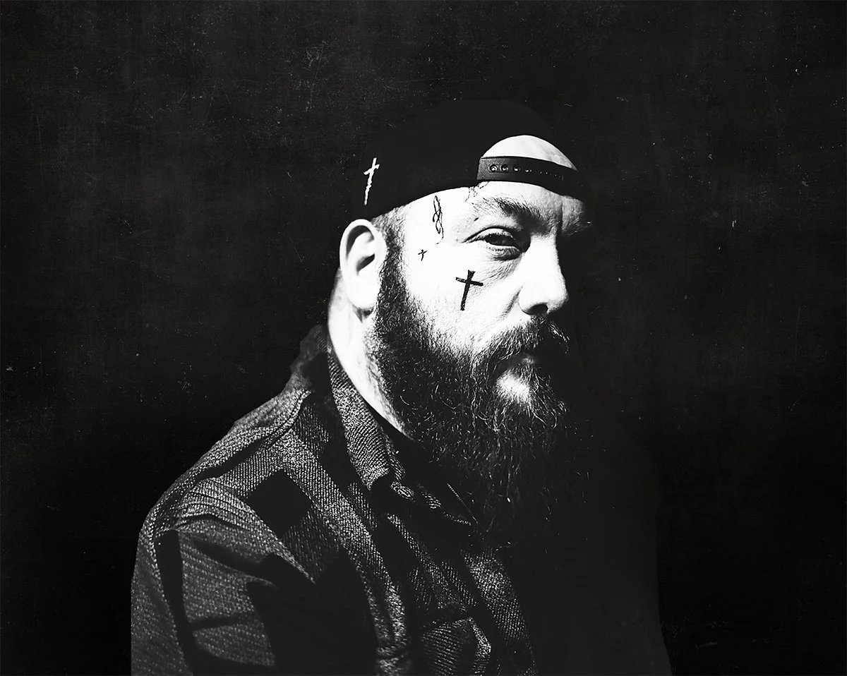 Black and white portrait of a bearded man with tattoos, wearing a baseball cap backward, against a dark textured background.