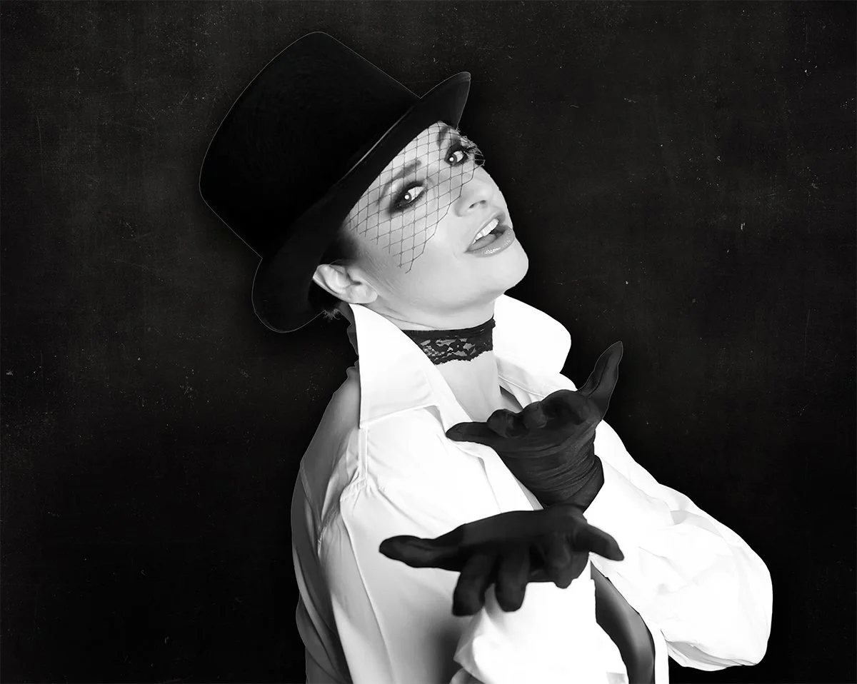 A woman dressed in theatrical costume with a top hat, lace veil, gloves, and a white shirt, posing against a black background.