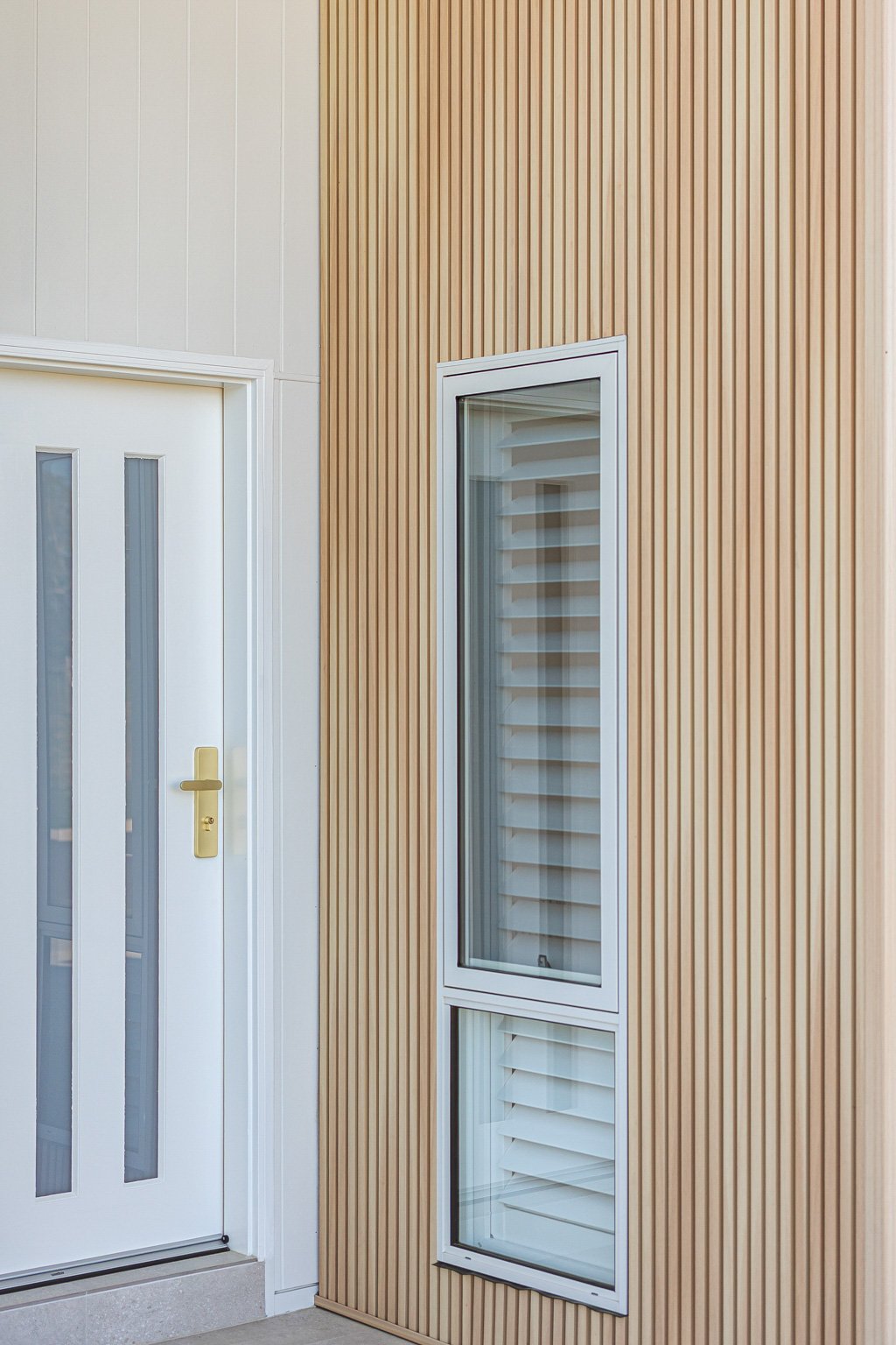 Exterior wall with vertical wood paneling, white door with narrow glass panels and a gold handle, and a tall, narrow window with louvers.