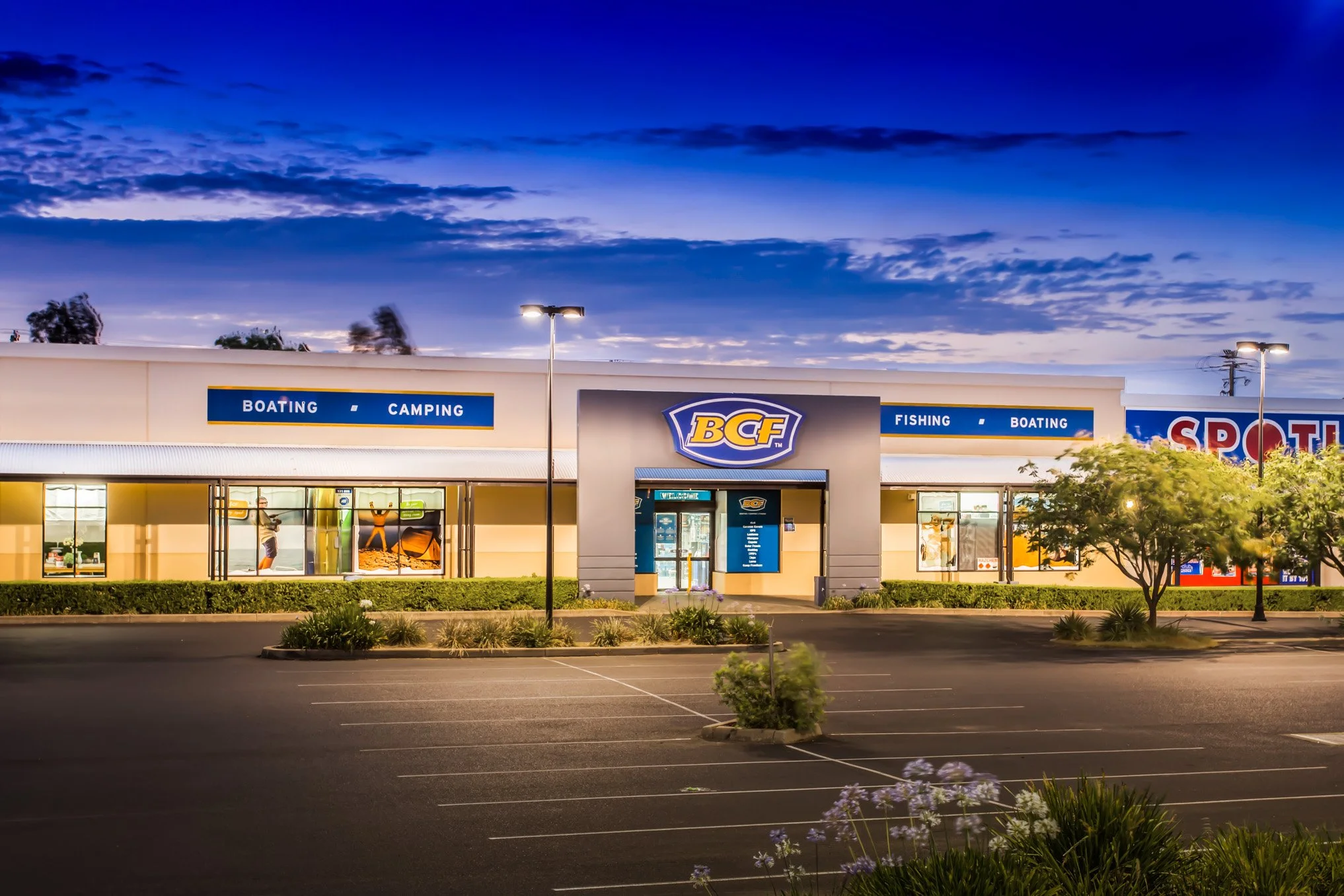 Exterior view of a sporting goods store at dusk with signage for fishing, boating, camping, and a large BCF logo, surrounded by a parking lot, trees, and streetlights.