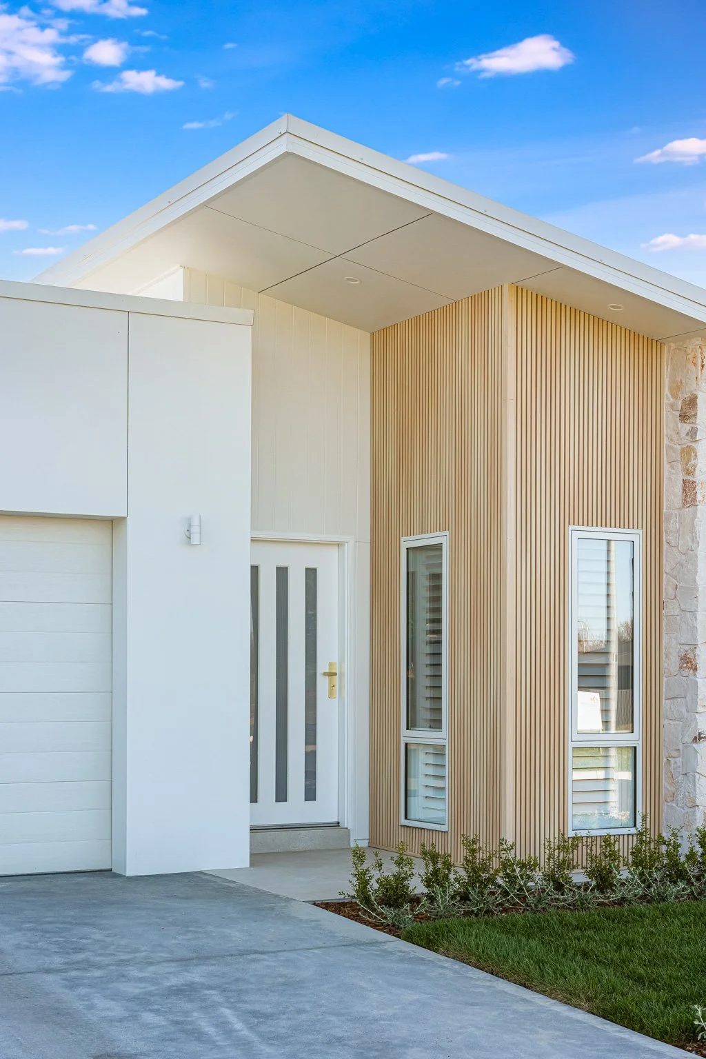 Modern house exterior with white garage door, front door with vertical glass panels, and two vertical windows with shutters, surrounded by small bushes and a concrete driveway.