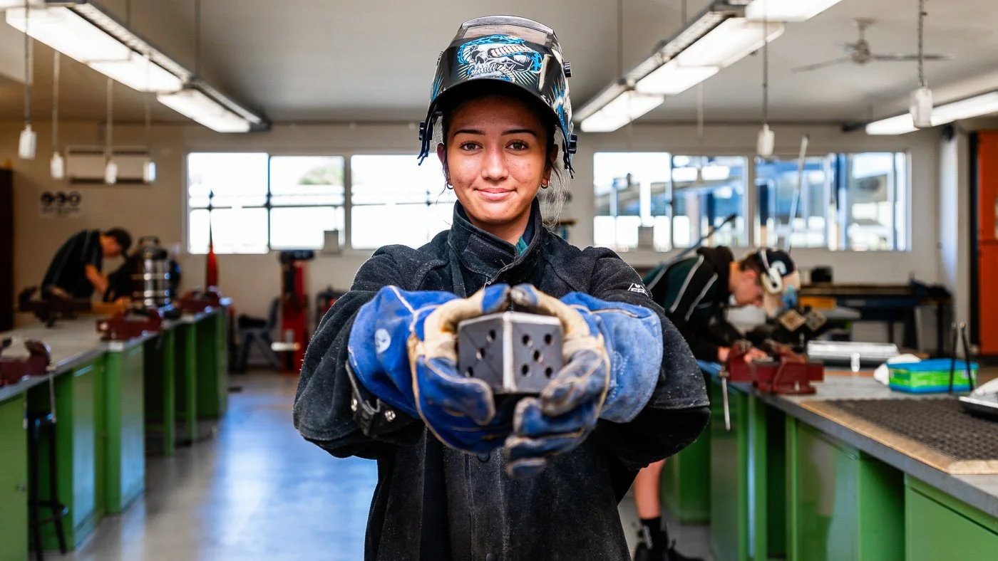 A young female welder in protective gear holding a metal piece in a workshop with other workers working in the background.