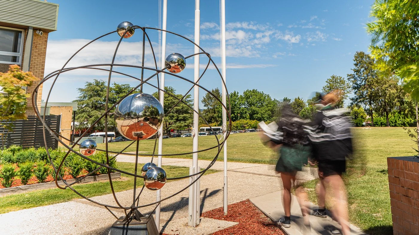 Metal sculpture resembling an atomic model with reflective spheres on a lawn, with two students walking past, some trees, a brick building, and a partly cloudy sky in the background.