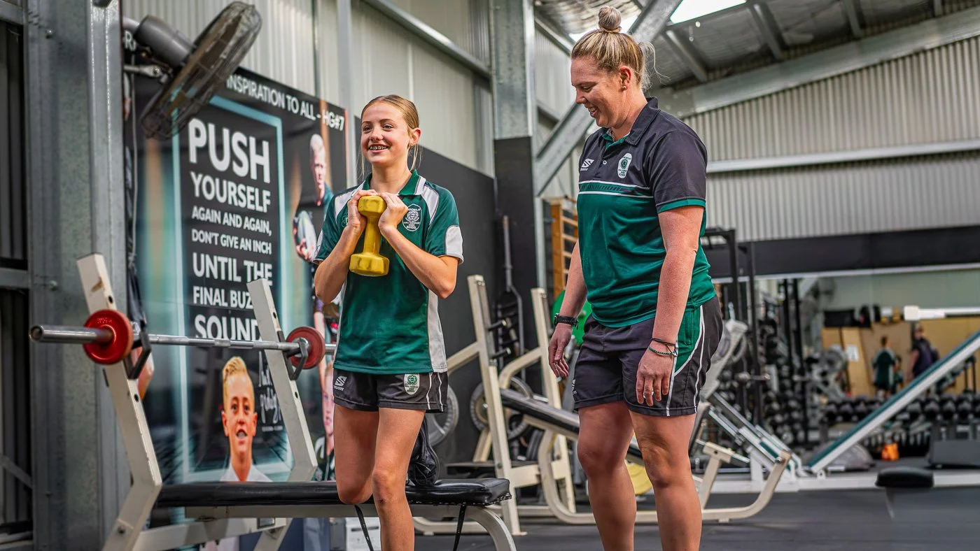 A young girl and an adult woman exercising together in a gym. The girl is holding a yellow dumbbell, smiling, and standing on one leg on a bench. The woman, wearing a black and green sports shirt, watches and smiles. There is gym equipment and a moti