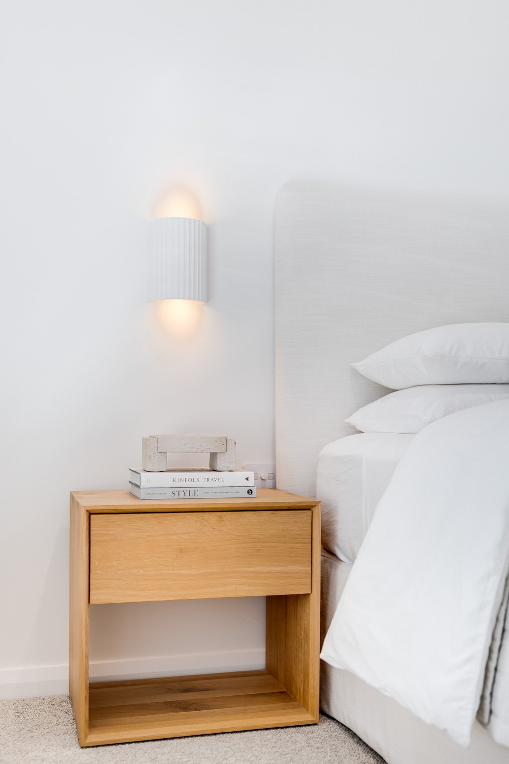 A minimalist bedroom corner with a wooden nightstand, a stack of books, a white wall-mounted light, and a bed with white bedding.