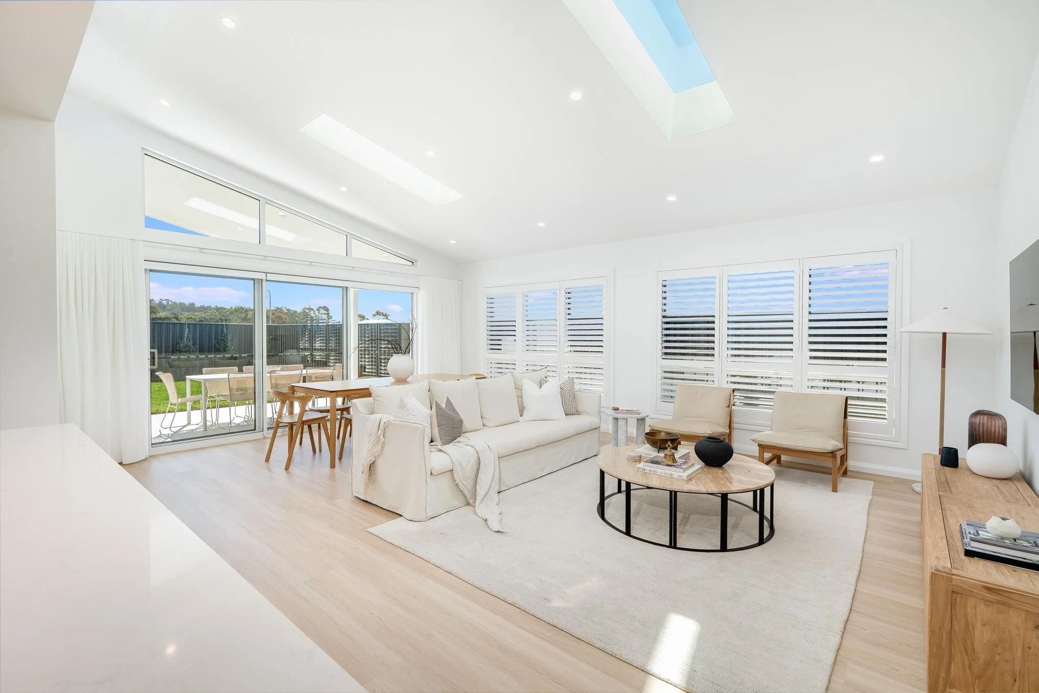 Bright and airy living room with a white sofa, beige chairs, and a round coffee table inside a modern home. Large windows with shutters, sliding glass door leading to outdoor patio, and a wooden sideboard with decorative objects.