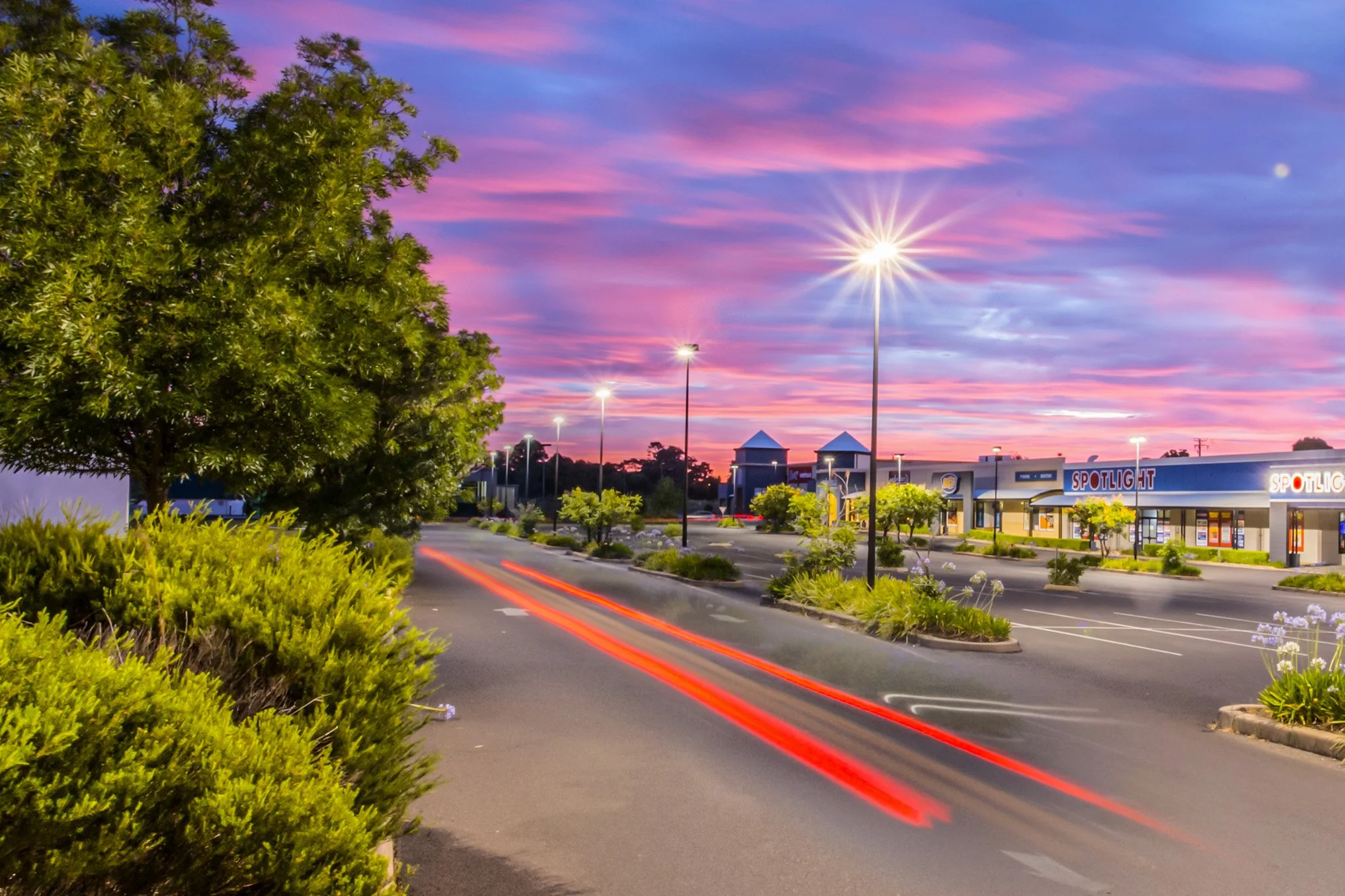 Empty shopping center parking lot at sunset with pink and purple sky, streetlights on, and light trails from passing vehicles.