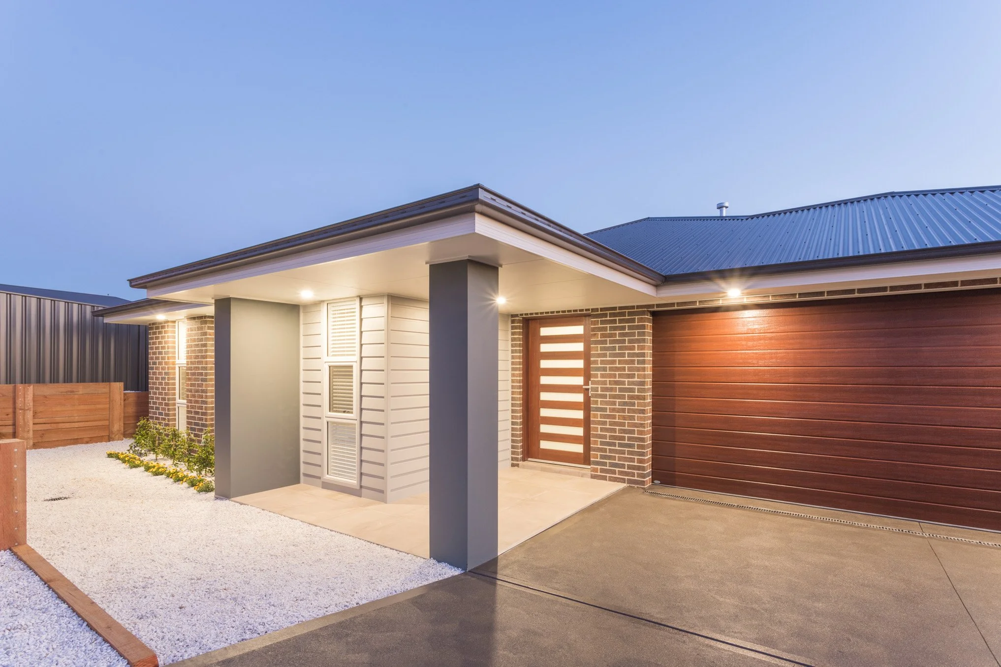 Modern house exterior with a wooden and brick entrance, driveway, and garden in the evening.