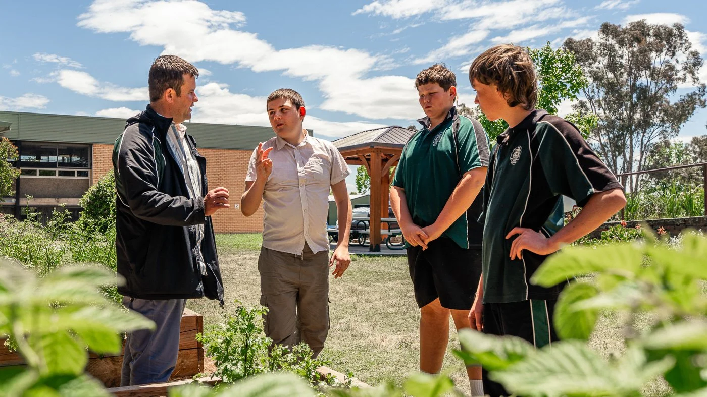 A teacher talking to four students outdoors at a school garden with a building, trees, and a blue sky in the background.