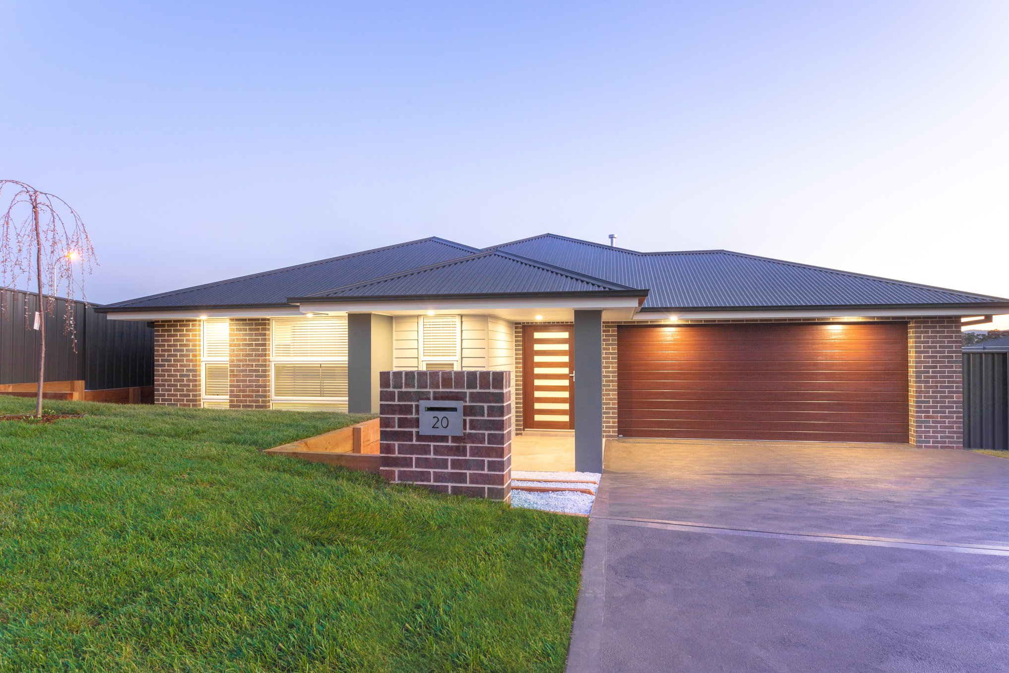 A modern, single-story house with a brick and siding exterior, illuminated by outdoor lights, featuring a two-car garage with a wooden door, a well-maintained green lawn, and a brick mailbox with the number 20, during twilight.