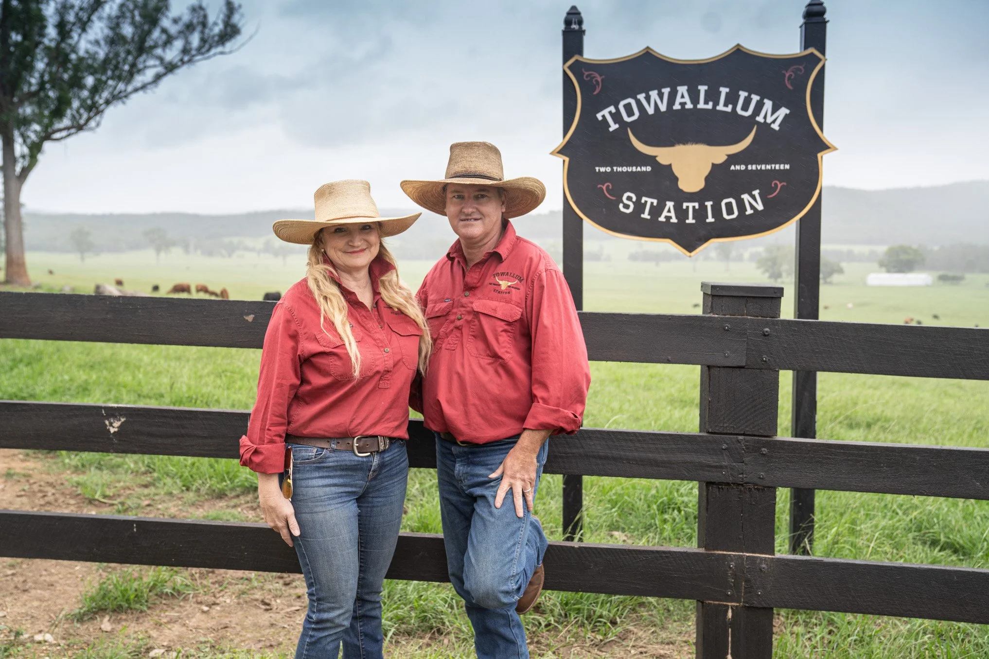 A man and woman standing outdoors behind a black wooden fence with a sign that reads 'Towallum Station' with a bull skull logo. Both are wearing red shirts and wide-brimmed hats, with a grassy field and cows in the background.