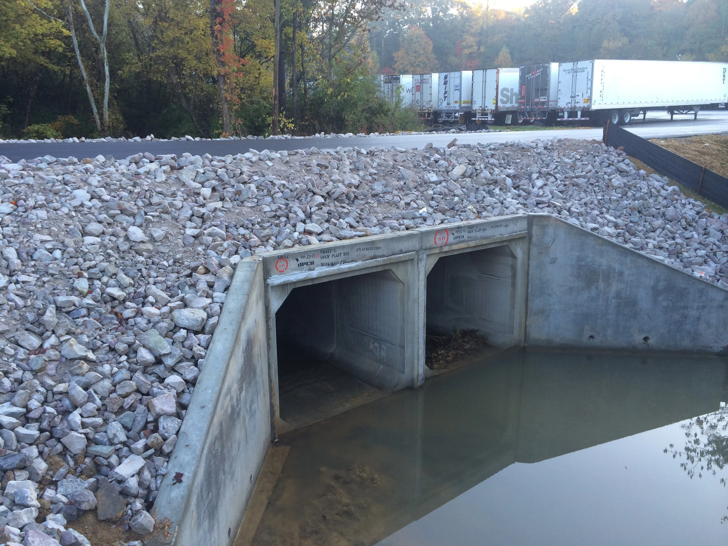 Construction site with a storm drainage culvert, rocks, water, and construction materials, with a highway and semi-truck trailers in the background.