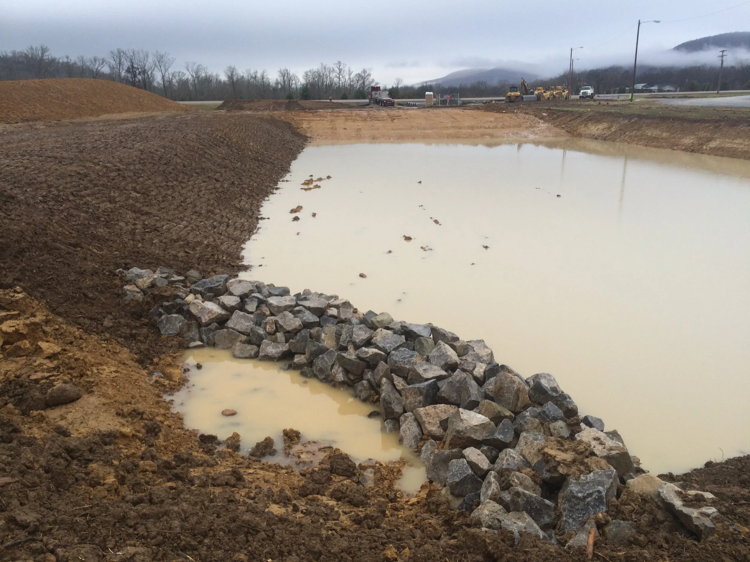 Construction site by a body of water with rocks along the edge, machinery, vehicles, and mountains in the background.