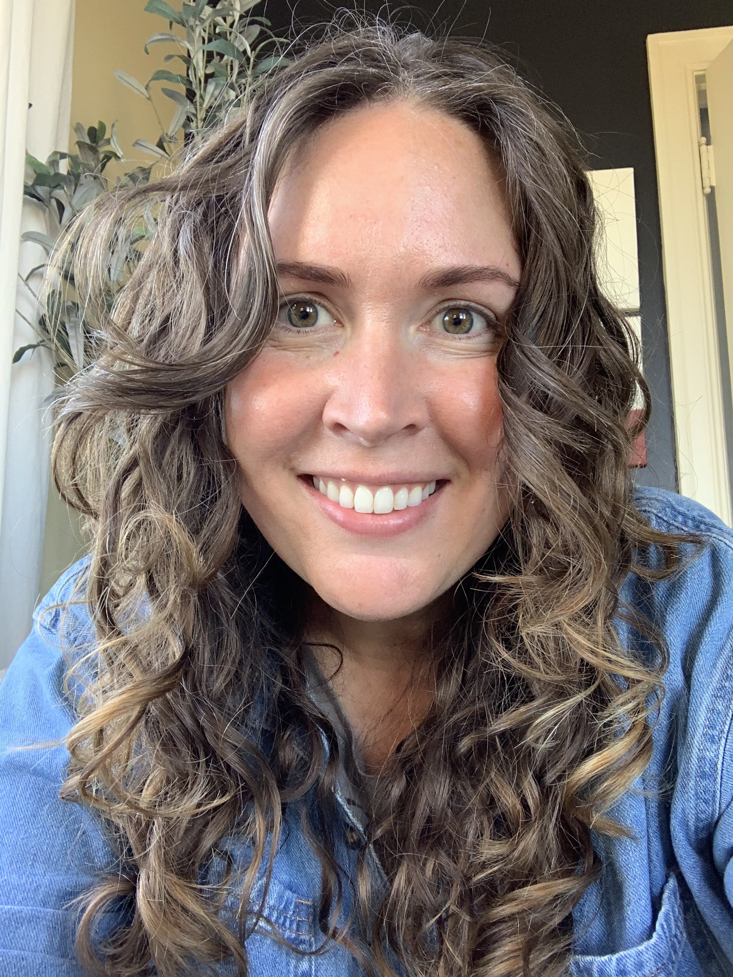 A photo of Morgan Shada, a woman with curly brown hair, smiling, wearing a denim shirt, standing indoors with a plant and a door visible in the background.