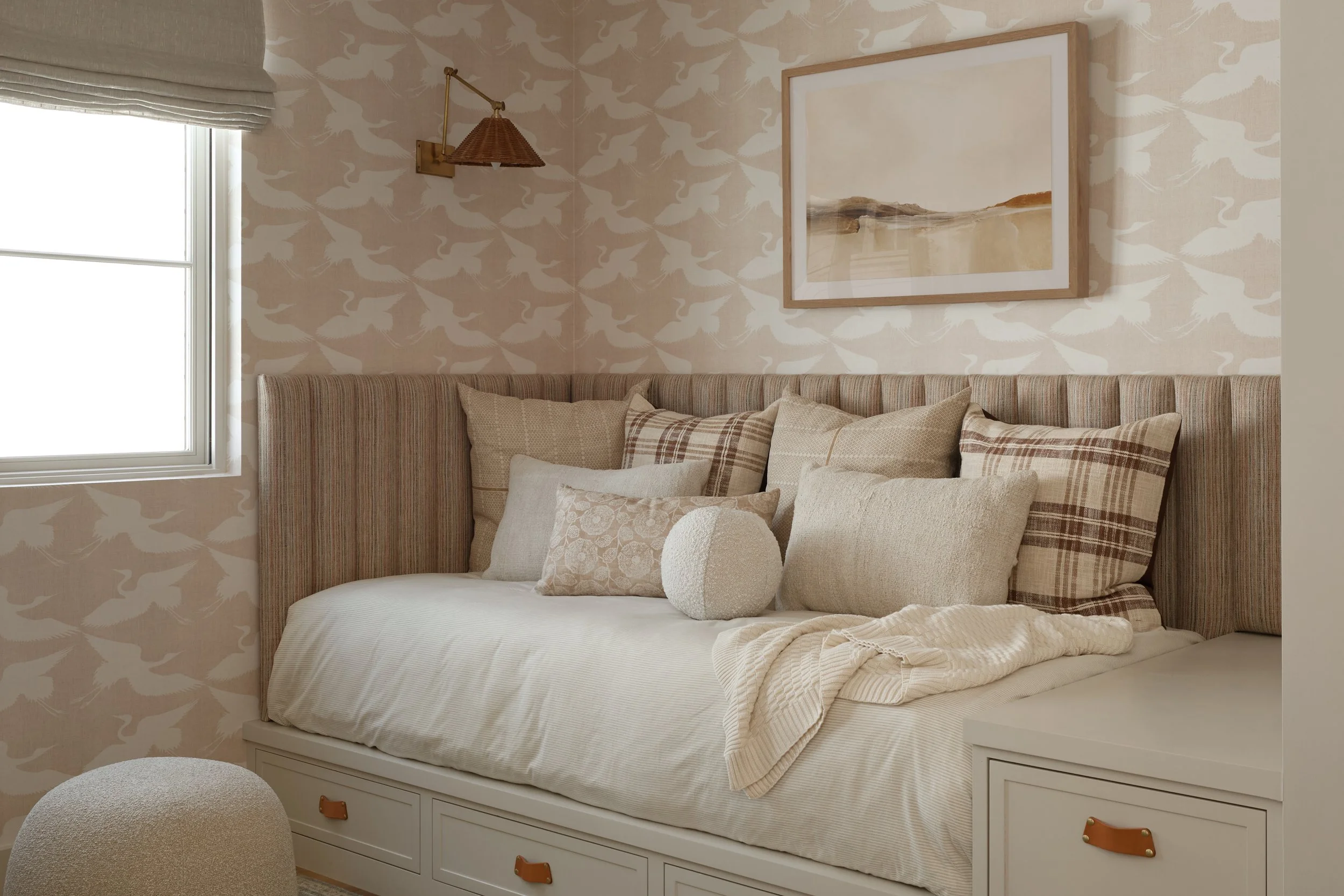 A bedroom with a daybed decorated with beige and plaid pillows, a window with a Roman shade, a wall-mounted light fixture, a landscape painting, and a beige wallpaper with a bird pattern.