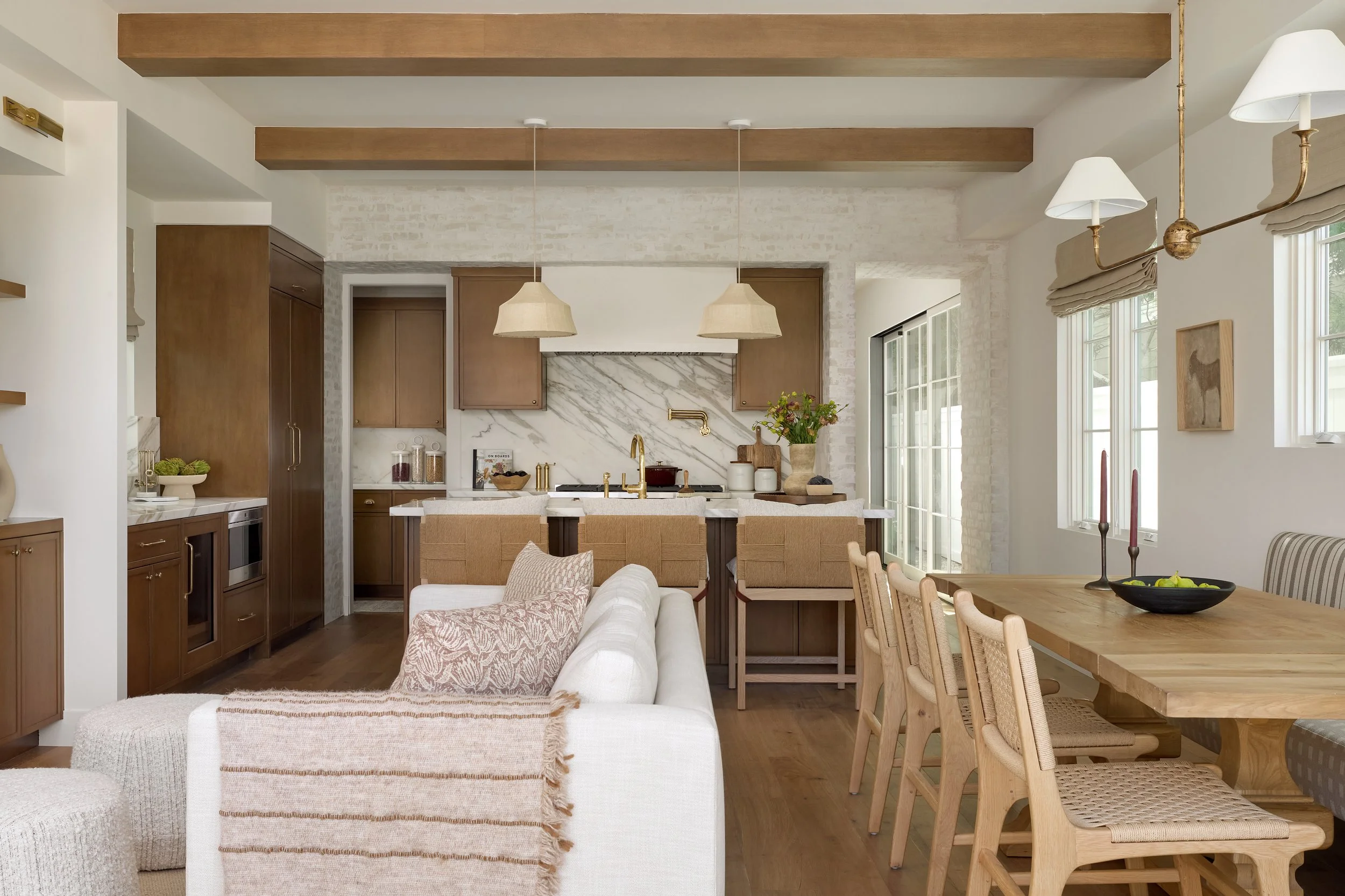 Open-concept living and dining area with a kitchen in the background, featuring wooden beams on the ceiling, white walls, a beige sofa with pillows, wooden dining table with wicker chairs, large windows bringing in natural light, and pendant lights o