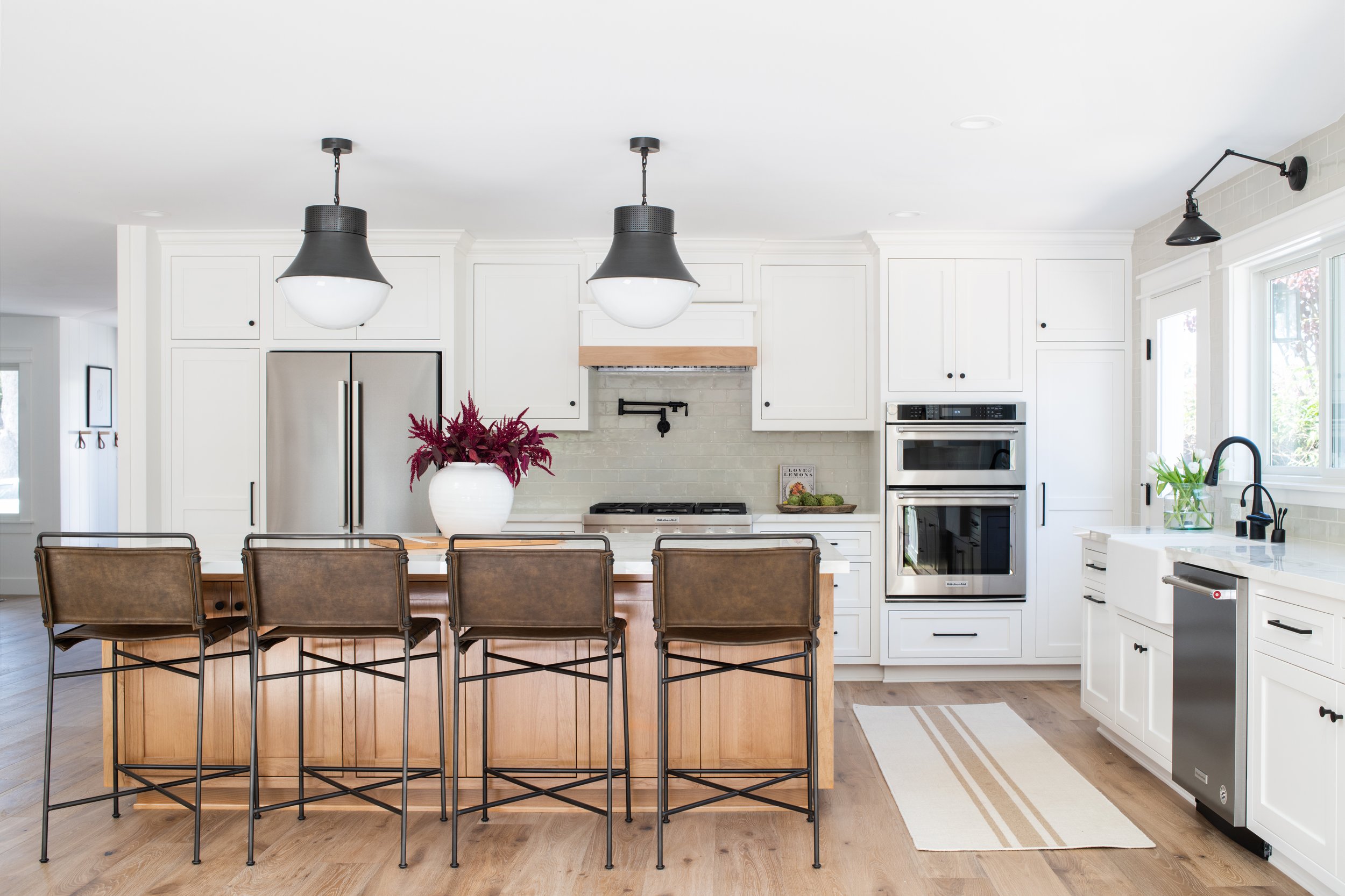 Modern white kitchen with wood accents, featuring a kitchen island with brown leather bar stools, a large white vase with flowers, stainless steel appliances, black pendant lights, and a beige striped rug. Los Angeles interior design studio.