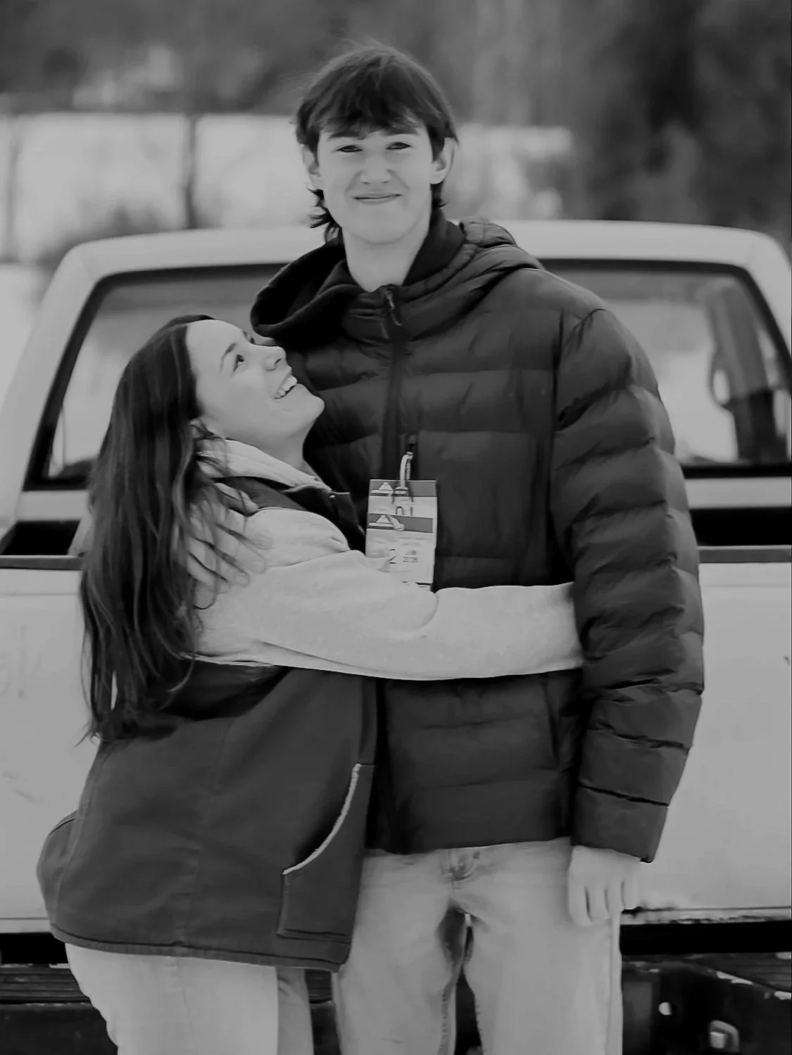 A young man and woman, smiling, hugging in front of a pickup truck. The woman is looking up at the man, wearing a jacket, and the man is dressed in a puffer jacket, with a name badge hanging around his neck.