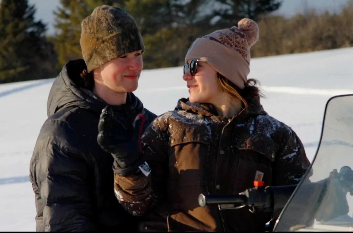 A young man and woman in winter clothing standing close together outdoors in a snow-covered landscape, smiling at each other.