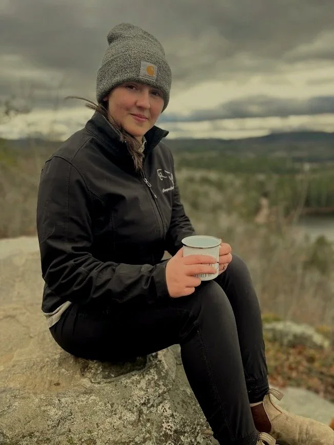 A young woman sitting on a large rock outdoors, wearing a gray beanie, black jacket, and black pants, holding a white mug, with a cloudy sky and landscape in the background.